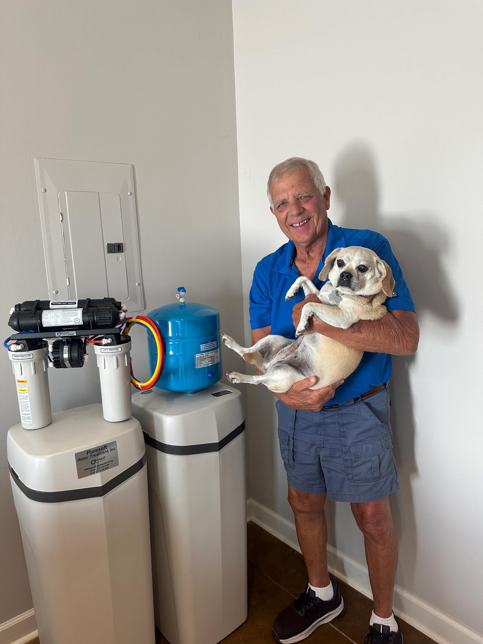 Man holding a small dog beside water filtration tanks in a utility room