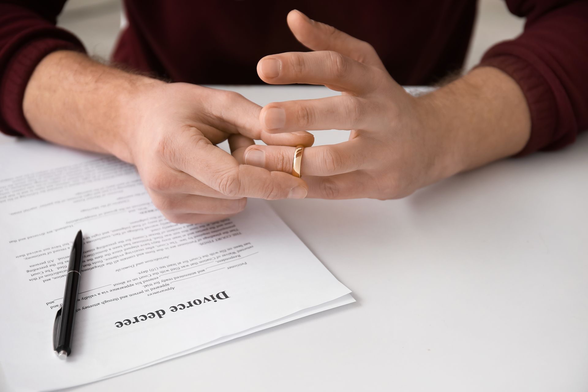 Person in a maroon sweater removing a wedding ring above a divorce decree paper and a black pen on a white table.