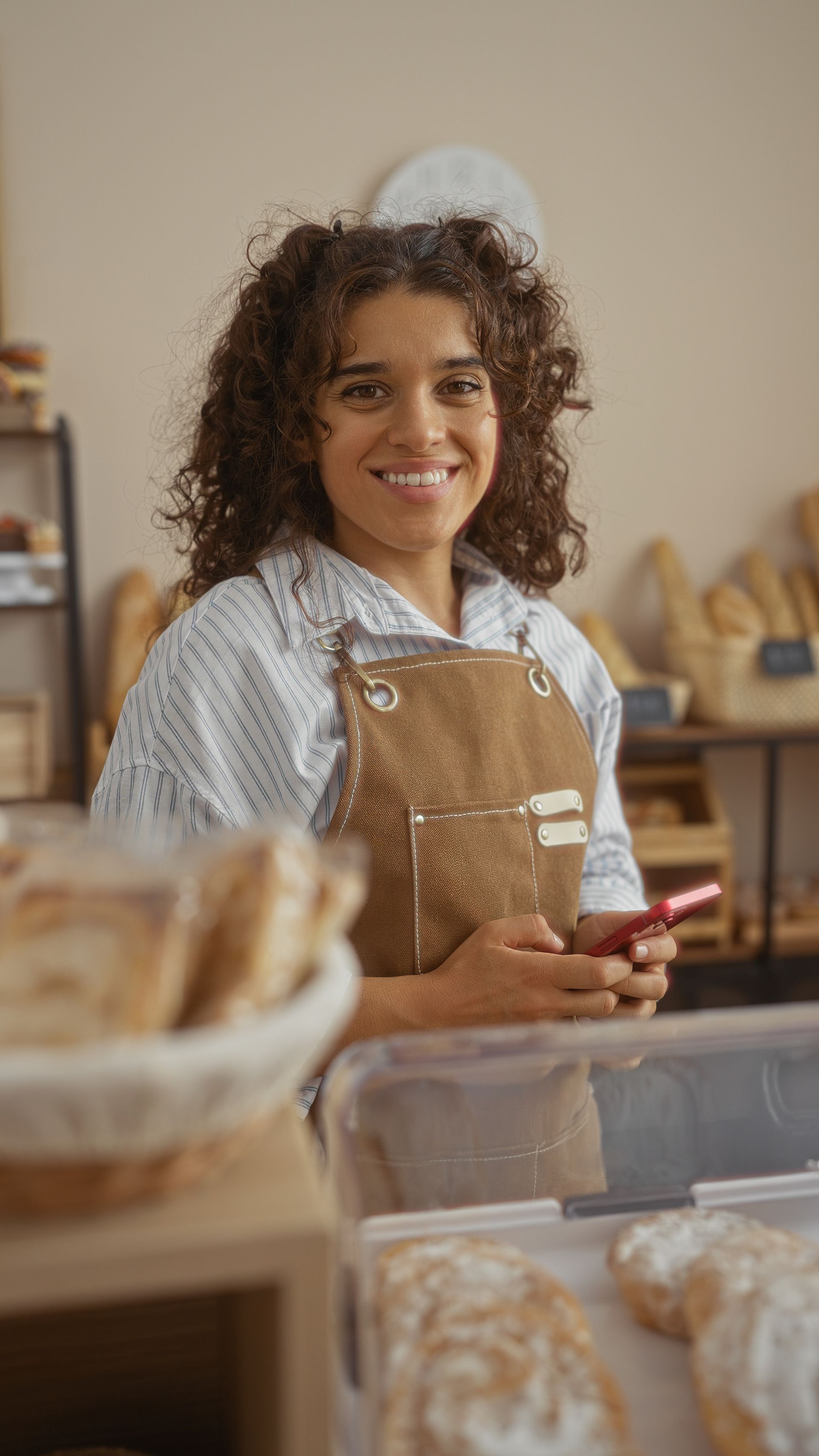 A woman is smiling while holding a cell phone in a bakery.