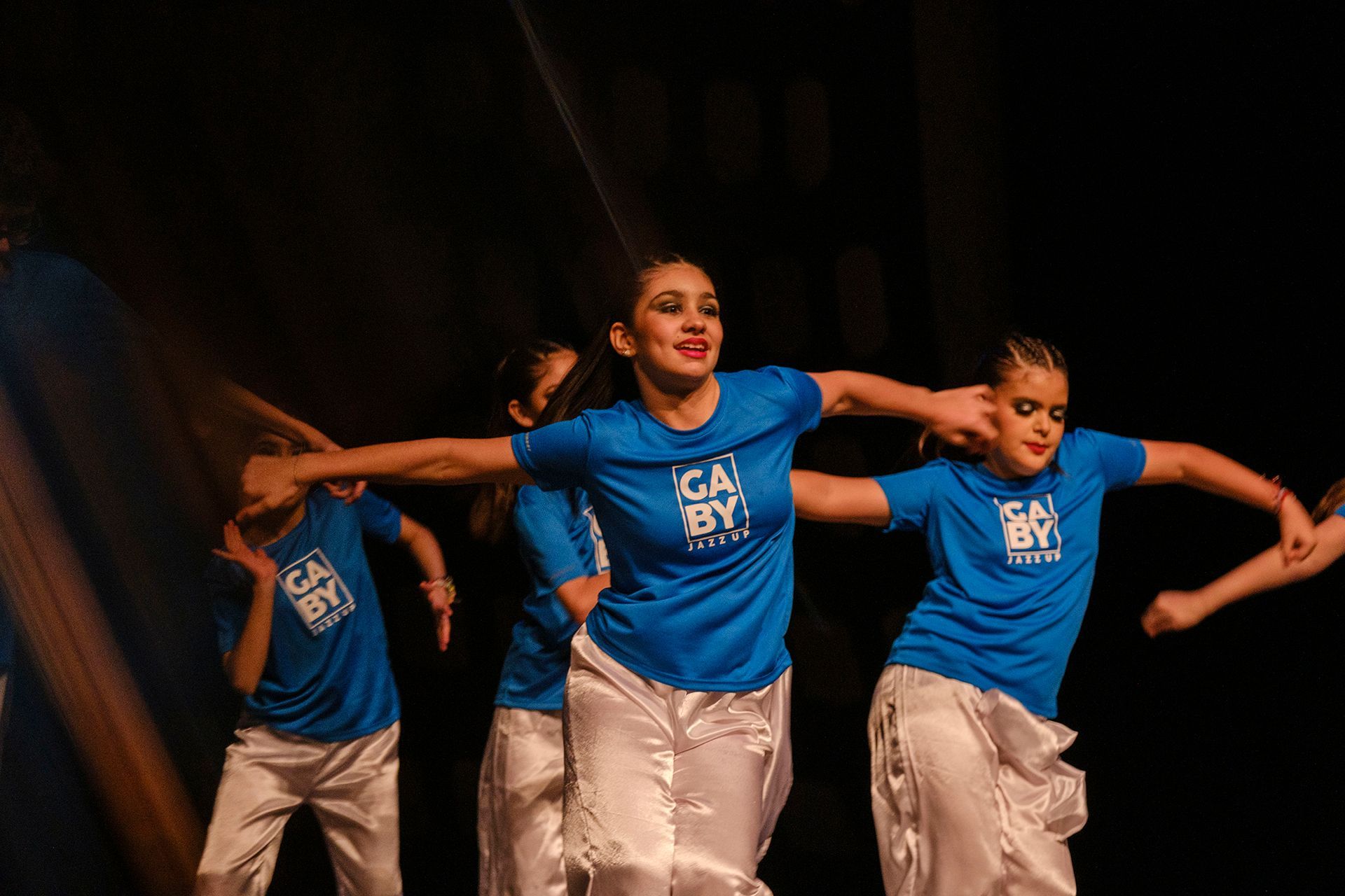 Bailarines con camisas azules y pantalones blancos actúan en el escenario. Tienen los brazos extendidos y expresiones sonrientes.