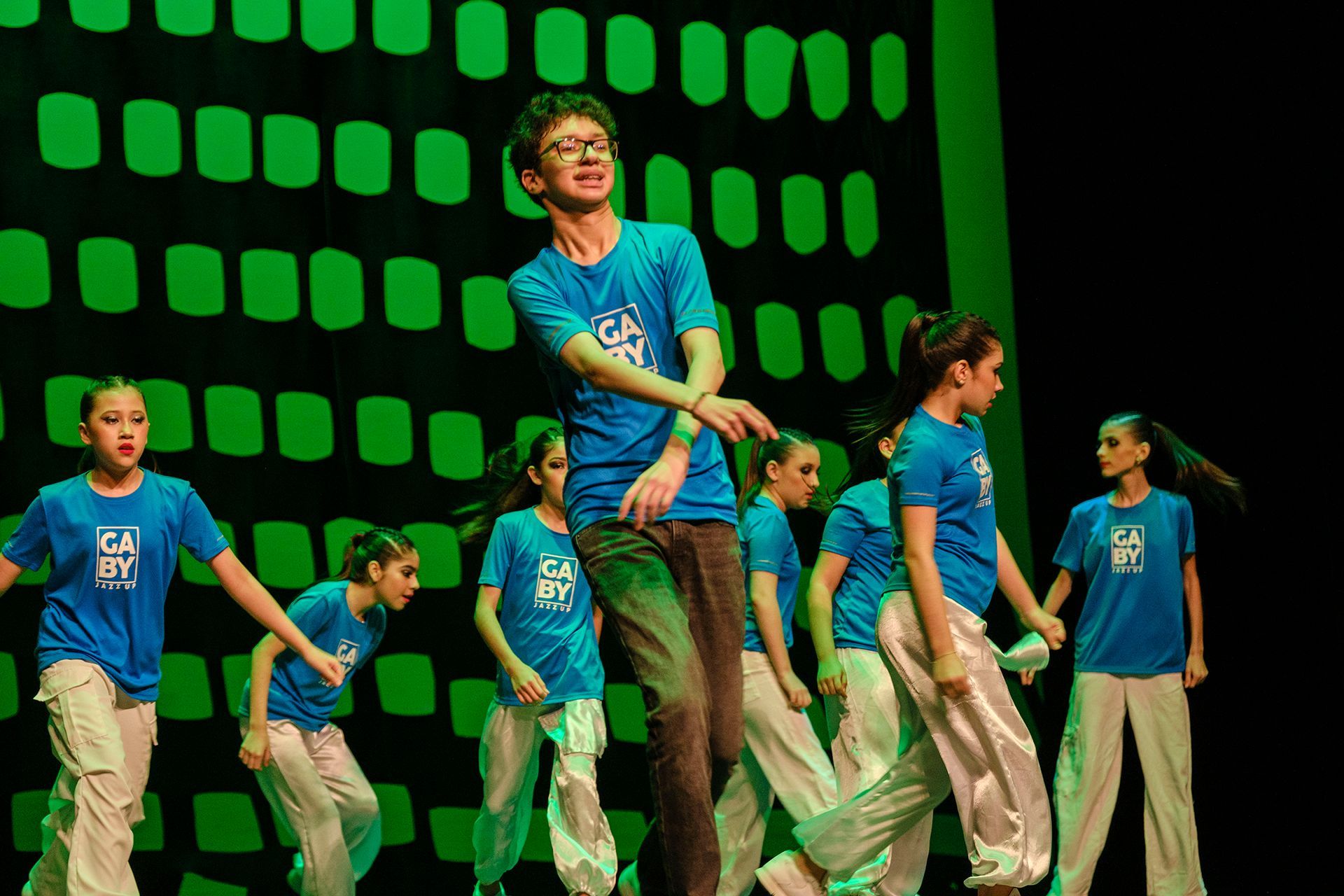 Bailarines con camisas azules y pantalones blancos actúan en el escenario con un fondo verde.