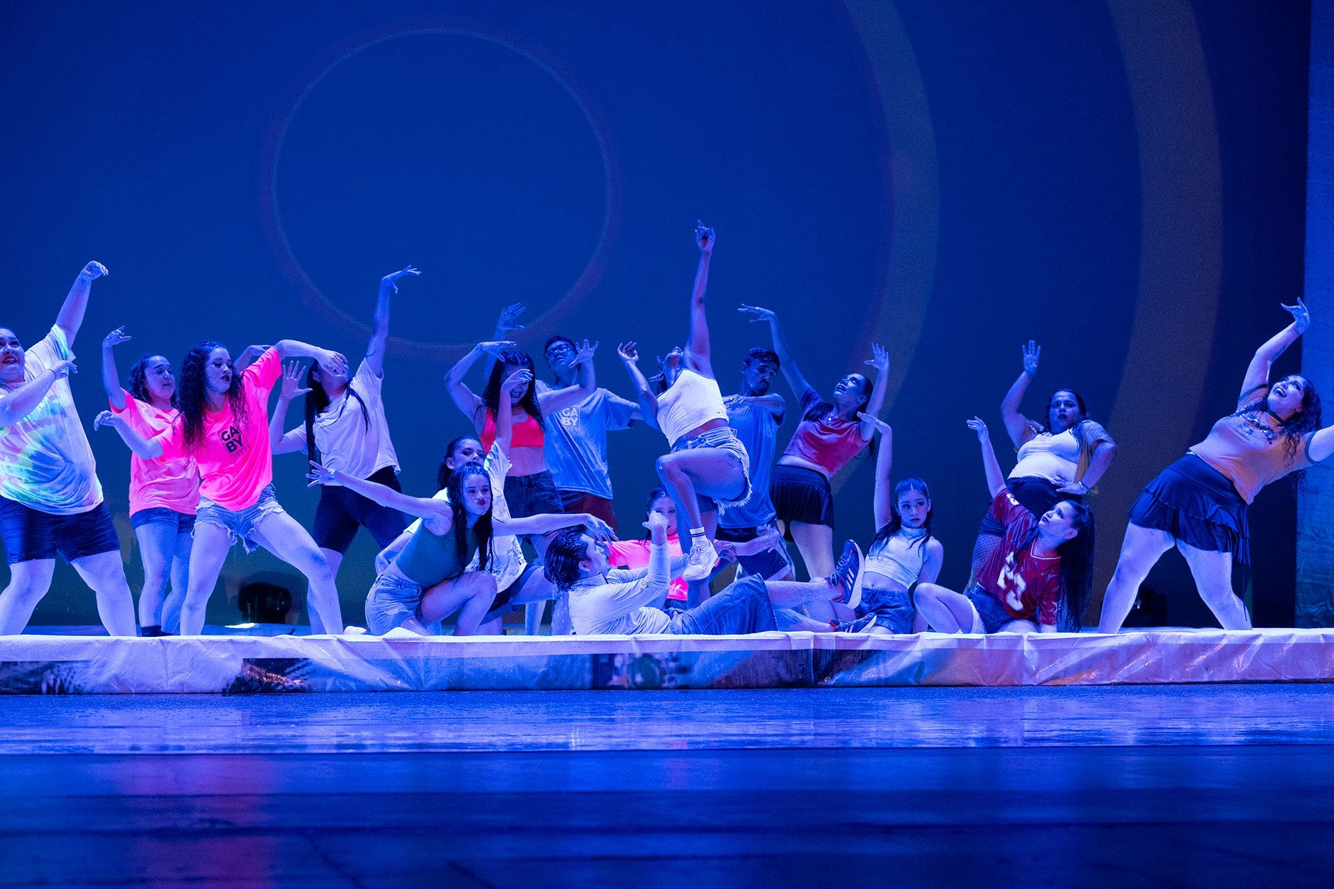 Bailarines en el escenario con los brazos en alto, iluminados por luz azul. Fondo colorido con círculos.