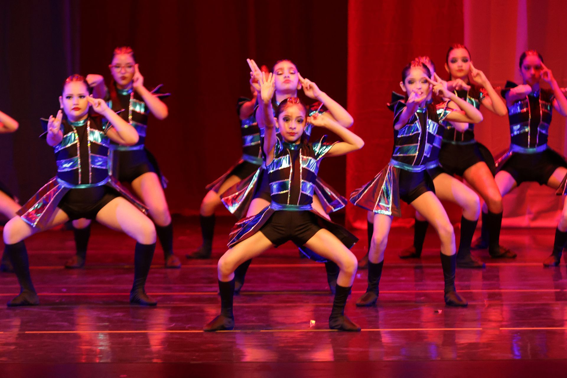 Bailarines con trajes negros y azules en el escenario, realizando una rutina sincronizada con los brazos en alto en un decorado escénico.