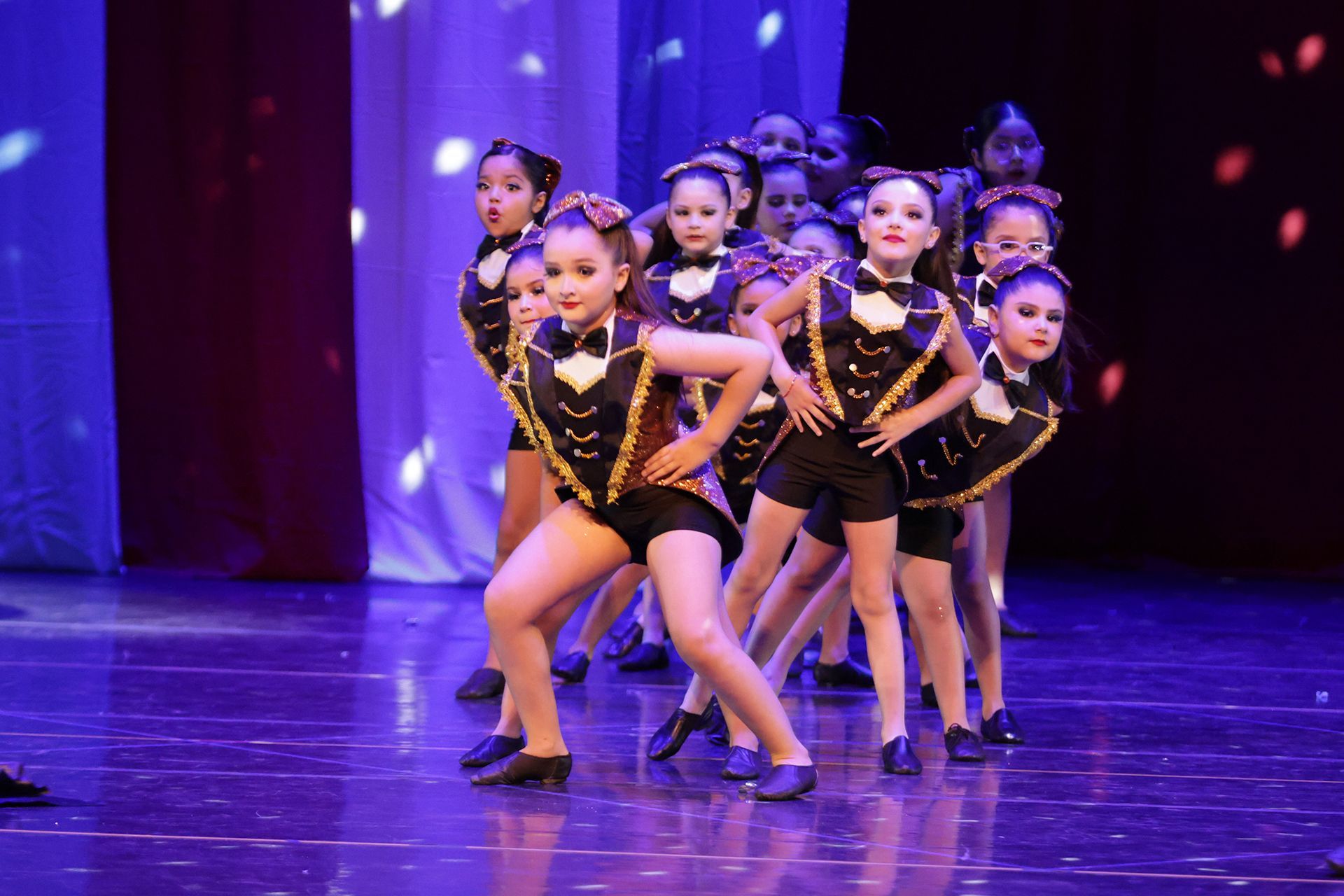 Jóvenes bailarinas con trajes negros y dorados en el escenario, posando con las manos en las caderas.