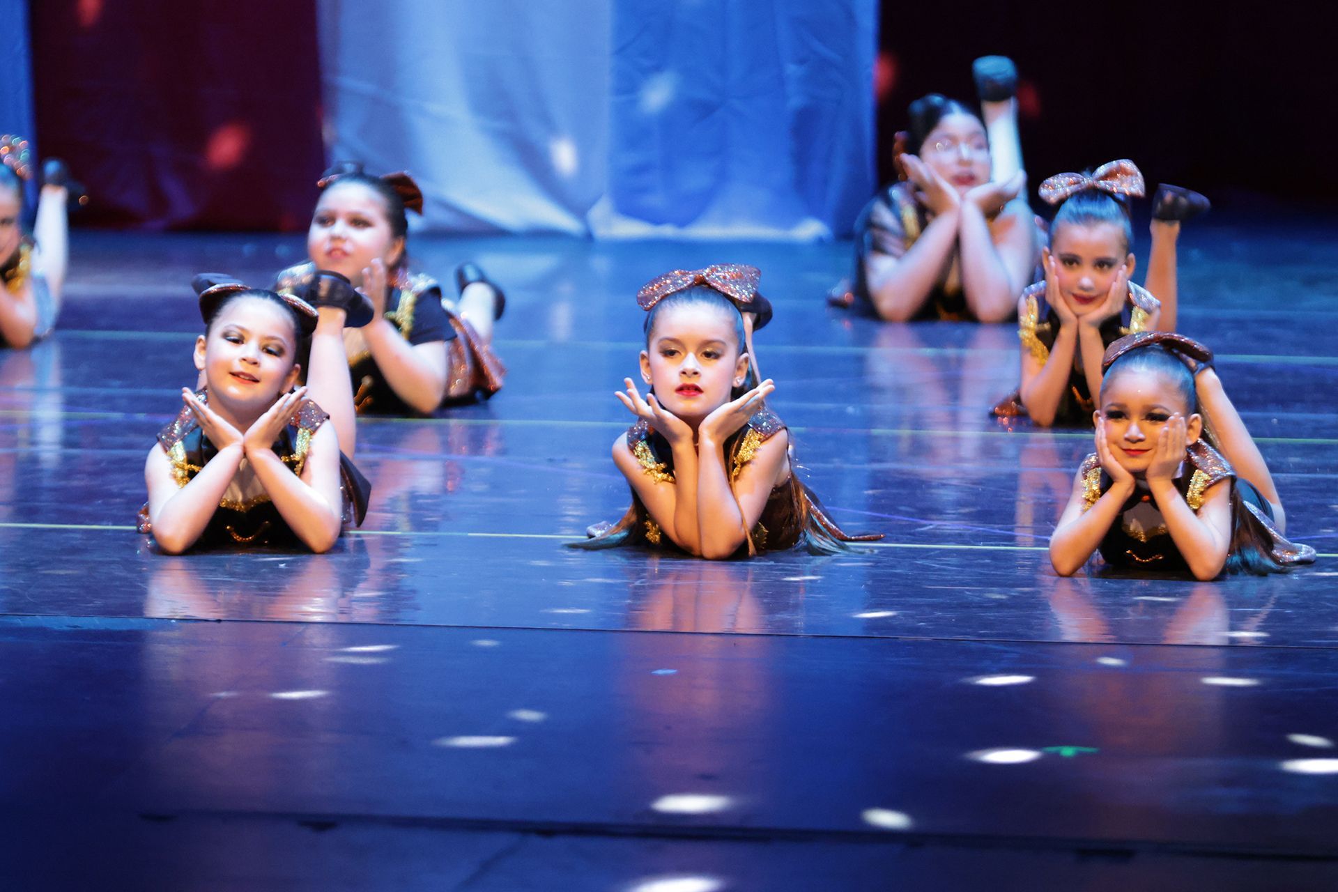 Bailarines con trajes negros y dorados en el escenario, posando con las manos bajo la barbilla, realizando una coreografía.