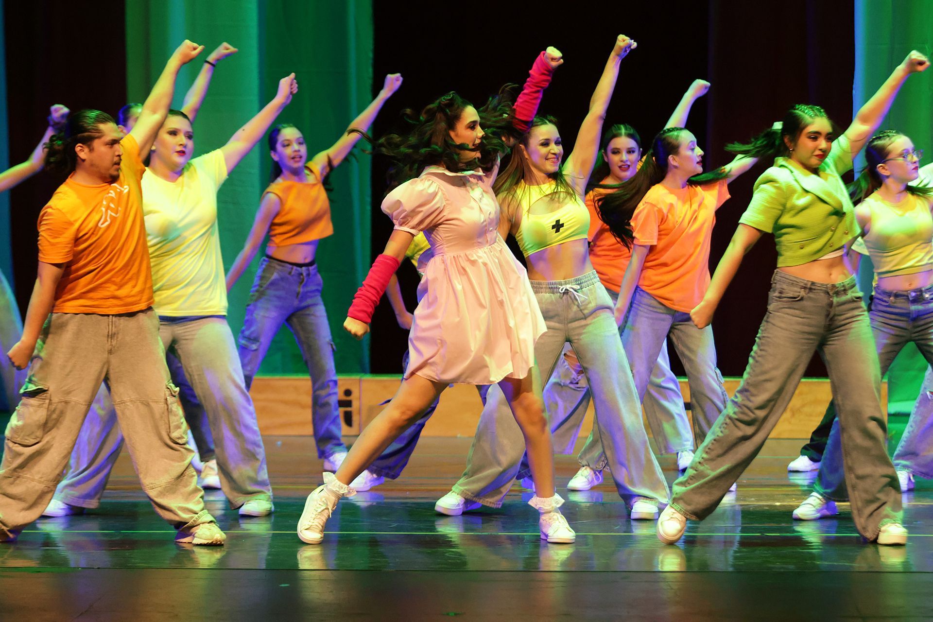 Bailarines en el escenario, con los brazos en alto, vestidos con camisetas coloridas y pantalones vaqueros. Iluminación verde, naranja y rosa.