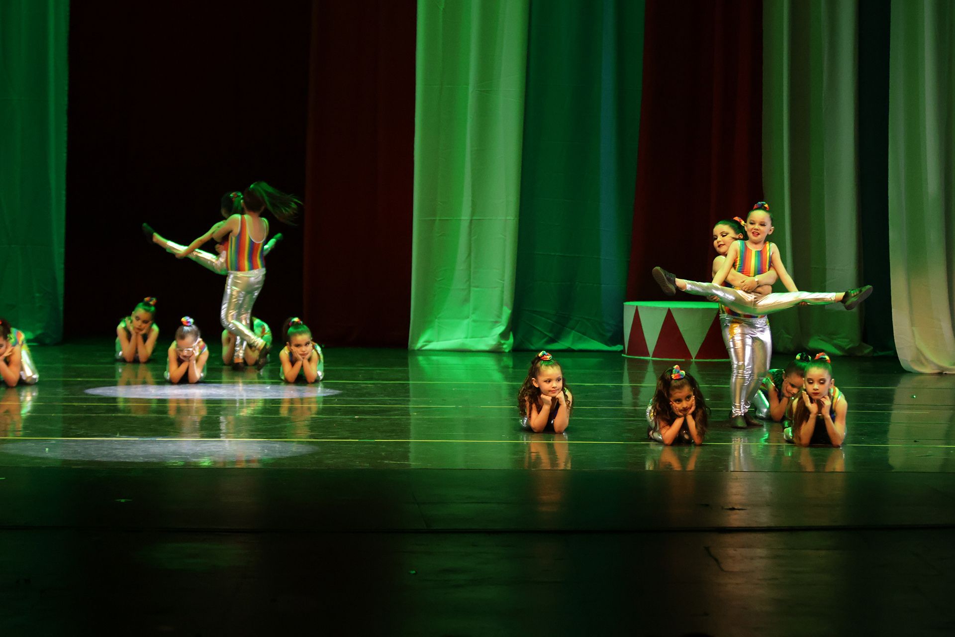 Bailarines en el escenario realizando movimientos acrobáticos, trajes verdes y plateados, fondo de cortinas rojas.