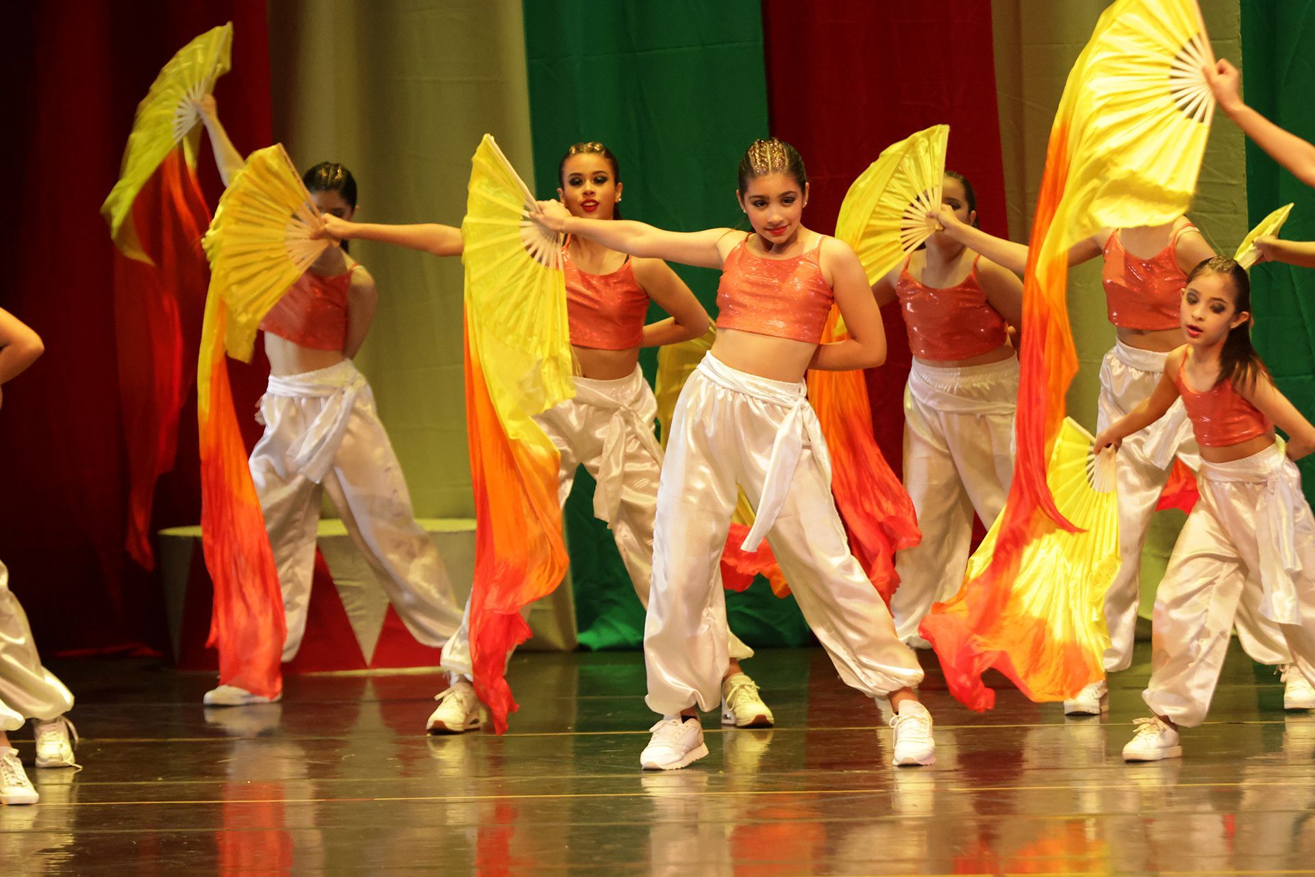 Bailarines en el escenario, sosteniendo abanicos amarillos y naranjas, vestidos con pantalones blancos y camisetas naranjas.