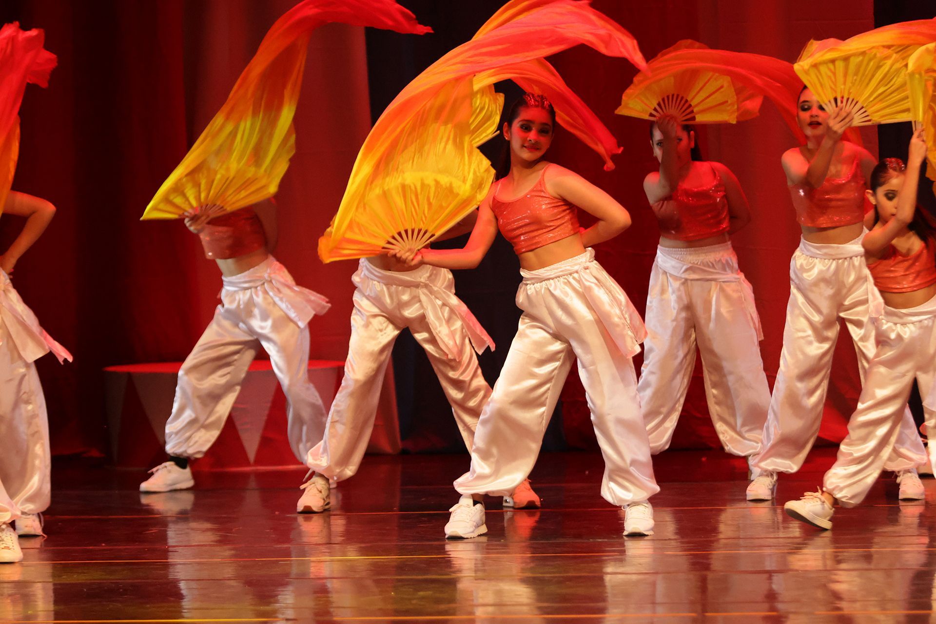 Bailarines con camisetas naranjas y pantalones blancos actúan con sus fans en un escenario con fondo rojo.