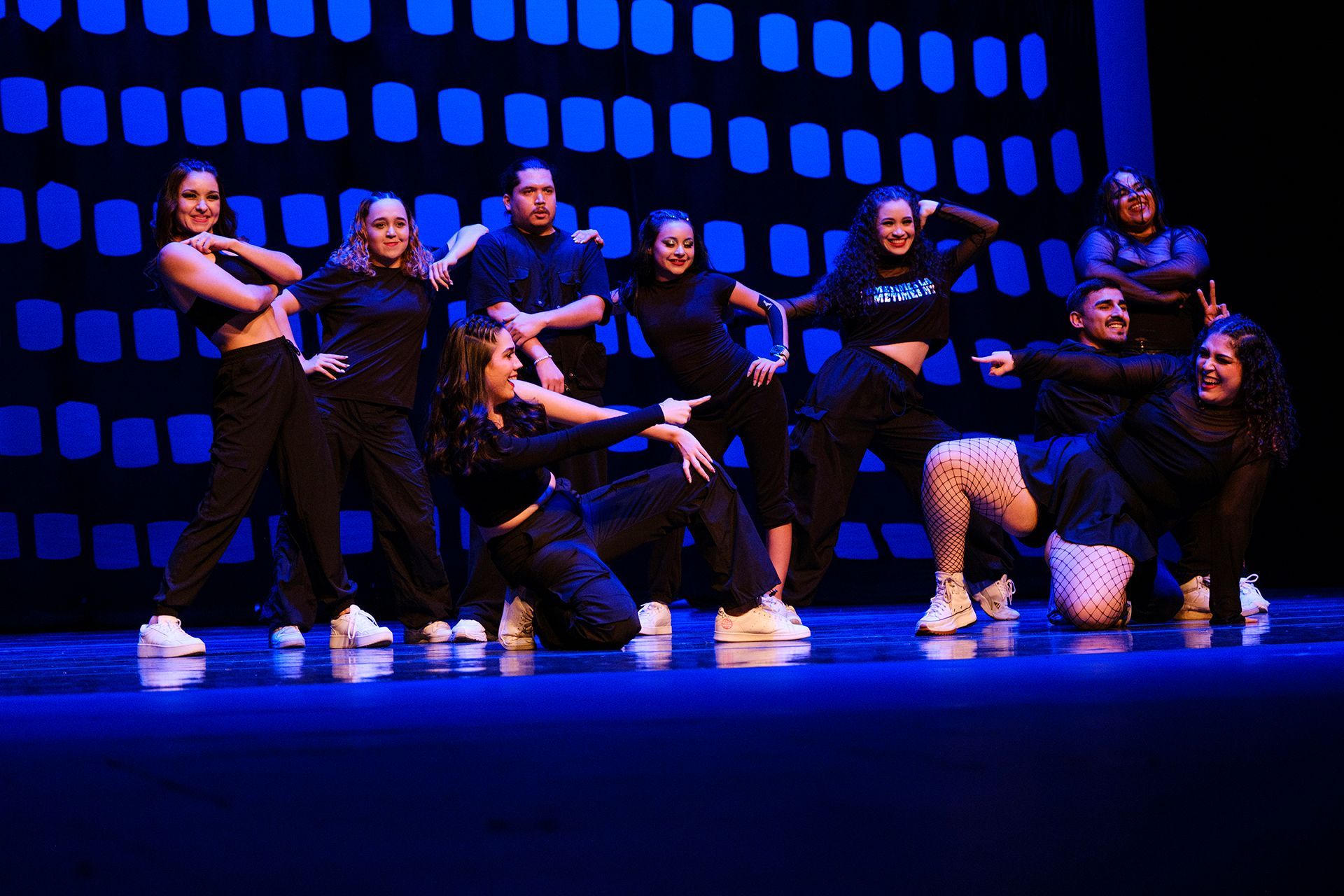 Un grupo de baile vestido de negro actúa en un escenario con un fondo que simula una ciudad.