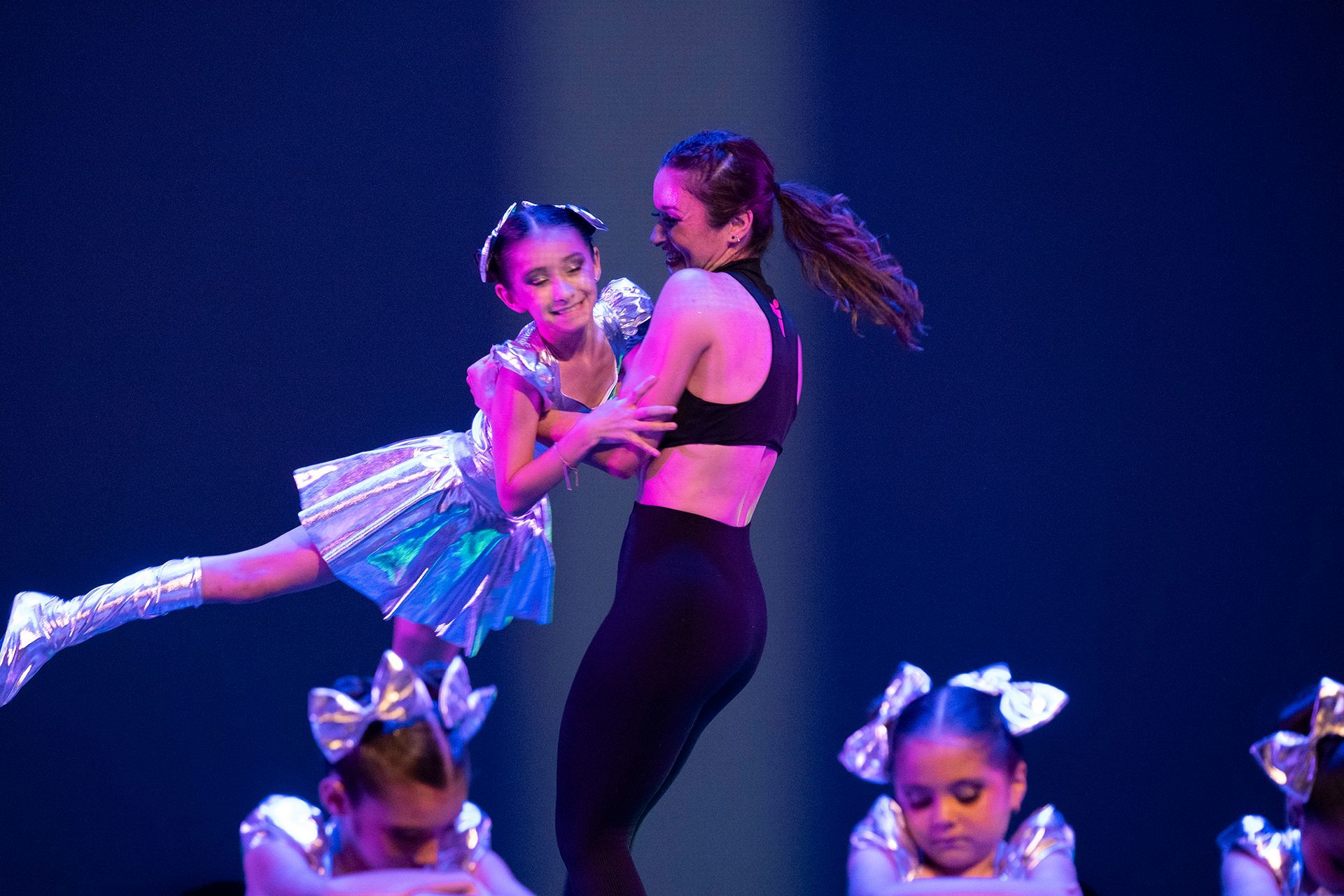 Bailarines actuando en el escenario; una niña pequeña con un traje azul es sostenida por una bailarina más alta vestida de negro.