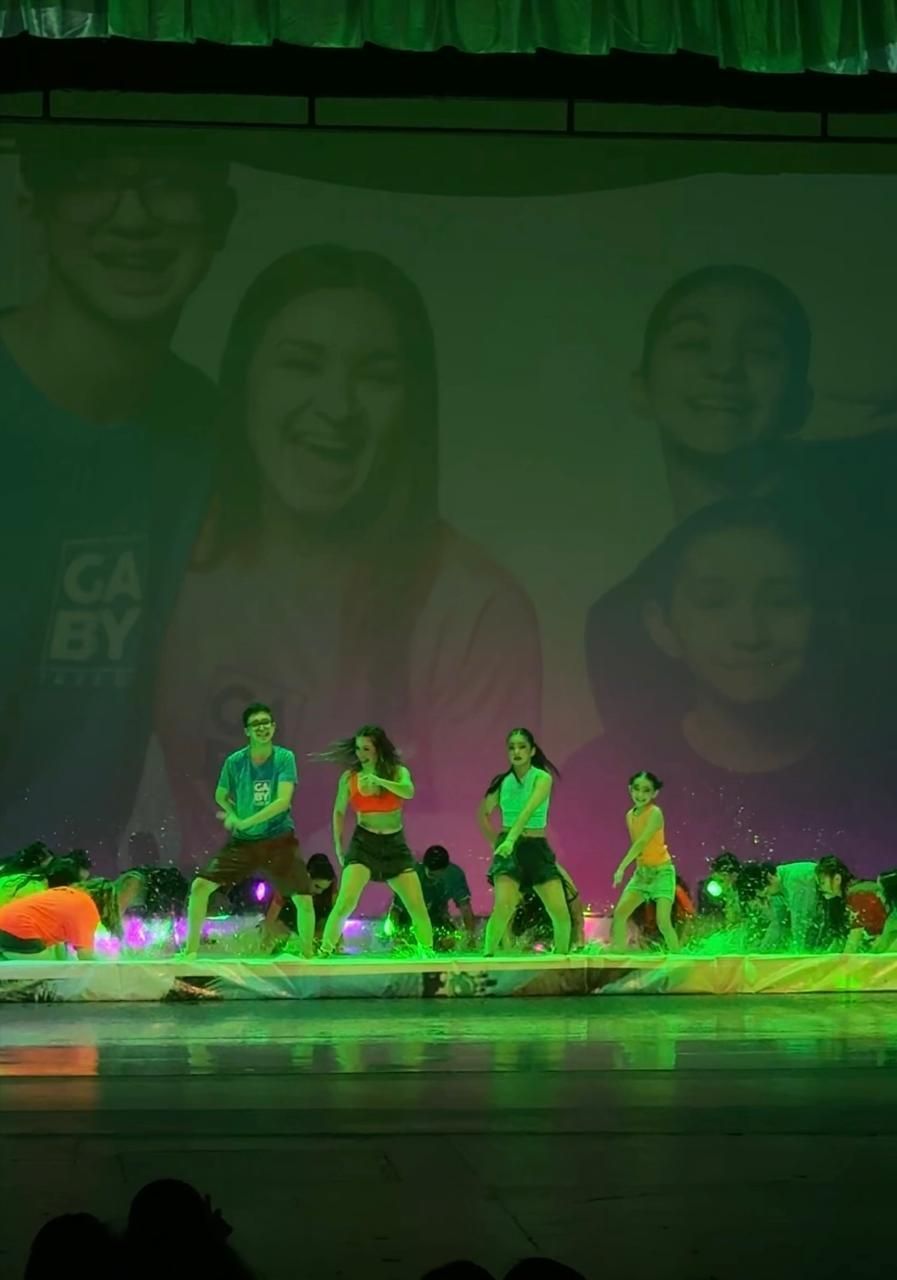 Los bailarines actúan en el escenario bajo una iluminación verde, con un fondo de rostros sonrientes. Visten trajes de colores brillantes.