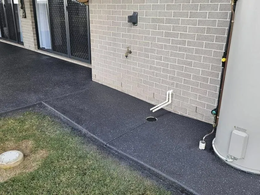 Black Paved Patio Next to a Grey Brick Wall, With a Water Tank and Plumbing — SRS Concreting In Gatton, QLD