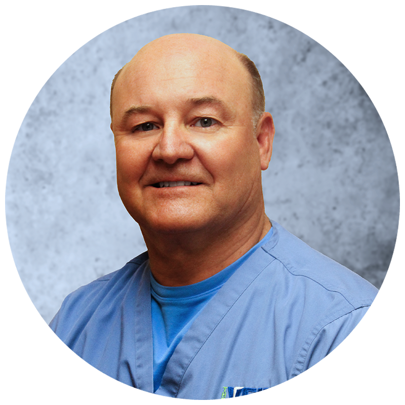 A bald man in a blue scrub top smiles for the camera