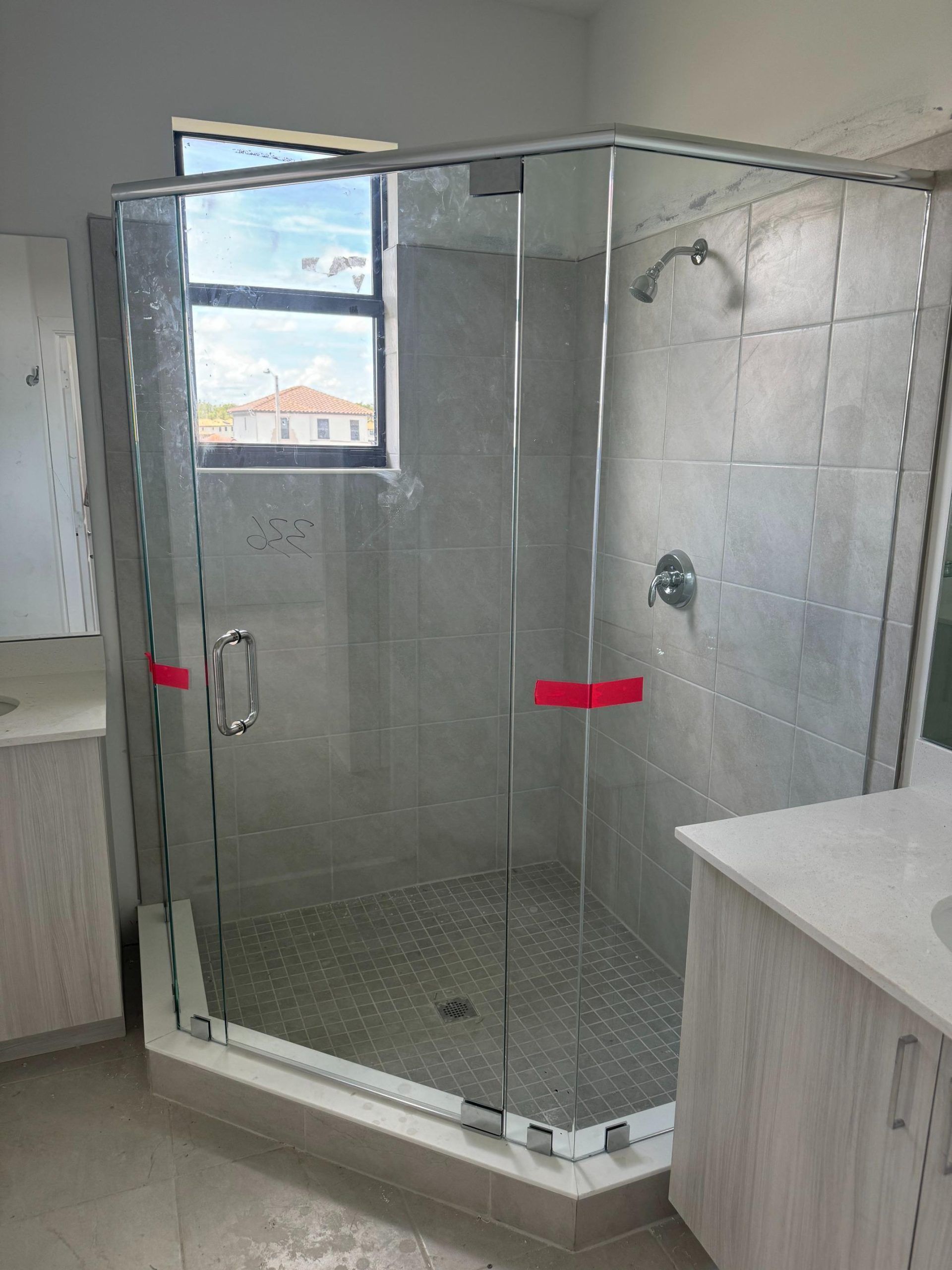 Glass shower enclosure in a bathroom with gray tiled walls and a small window.