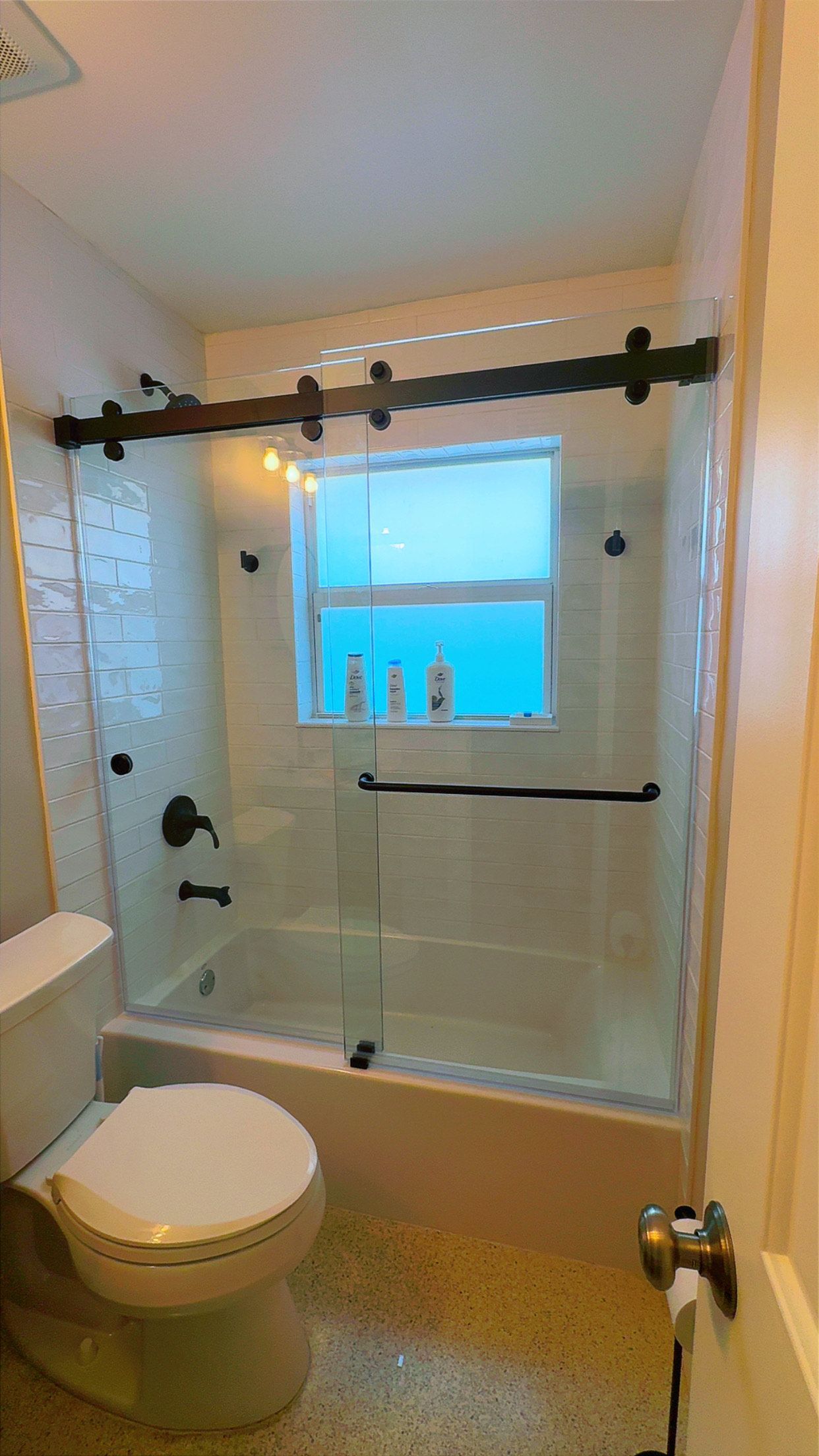 Bathroom with a glass shower door, toilet, and window. Black hardware and white tile are visible.
