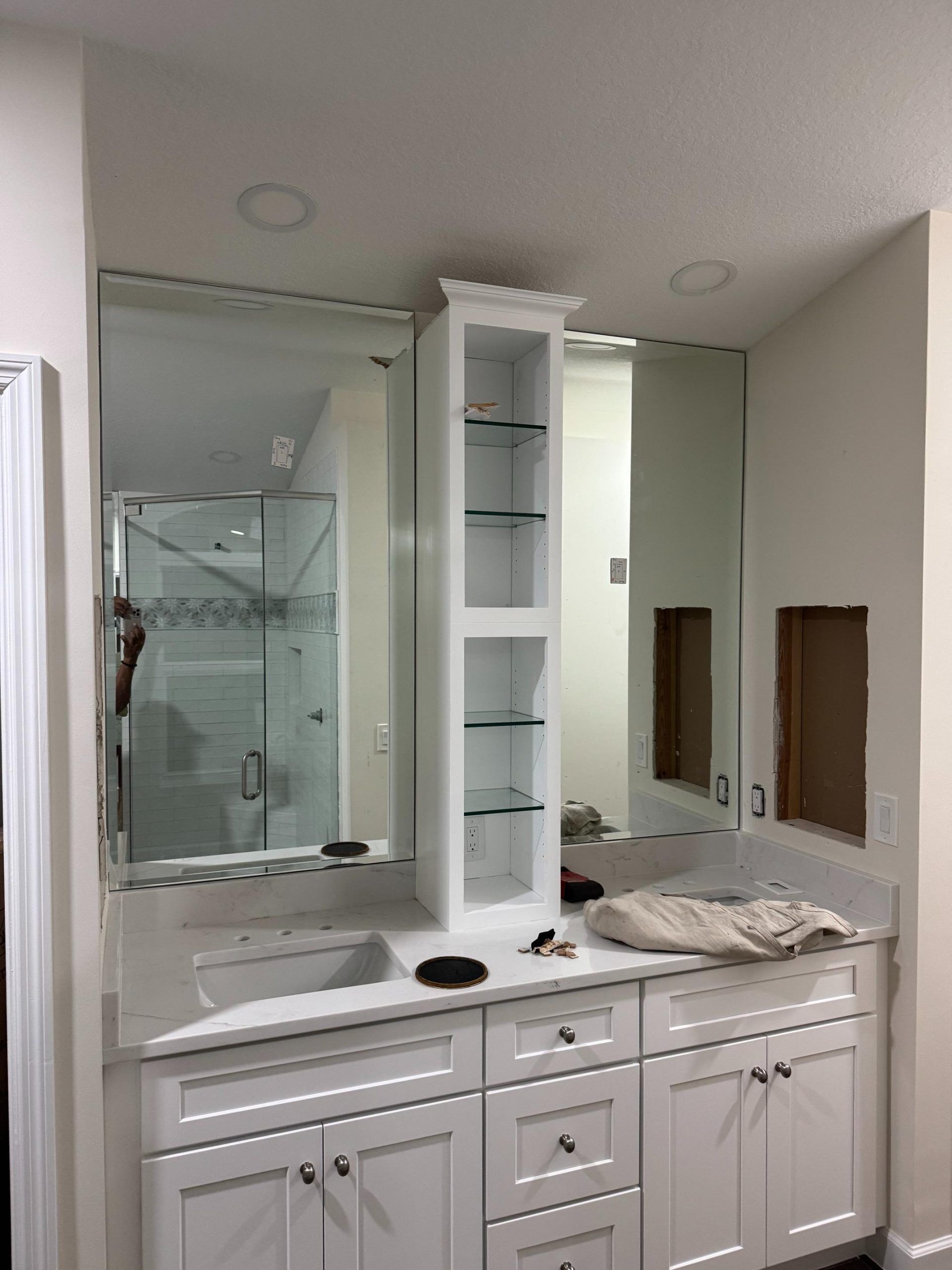 White bathroom vanity with mirrors and a central cabinet; a sink, and a shower are visible.