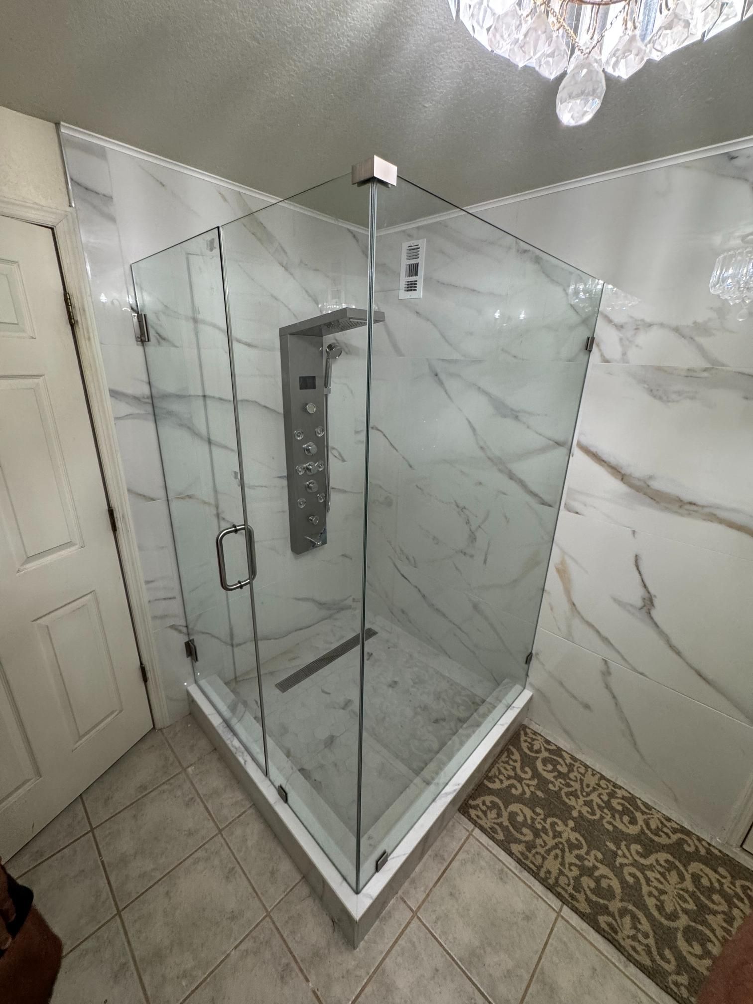 A modern glass shower enclosure with marble-look tile walls, a rain shower system, and a small decorative rug.