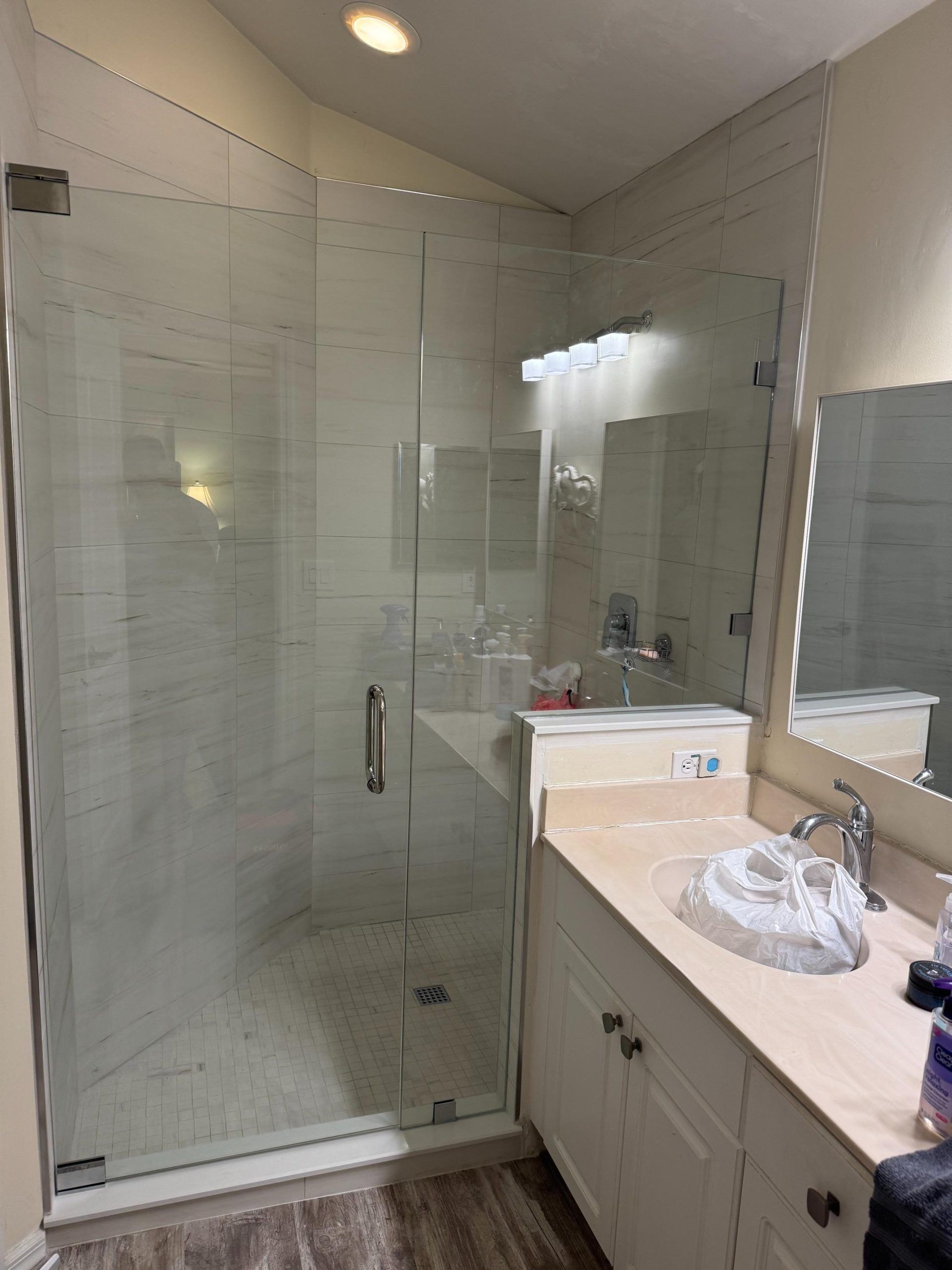 Glass shower enclosure with white tiled walls, next to a white vanity with a tan countertop and a mirror.