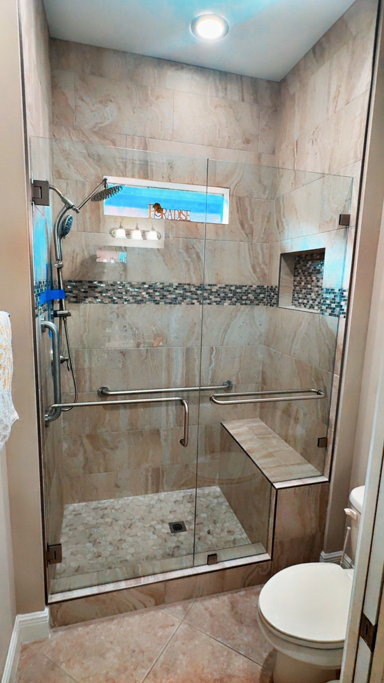 Shower with tan tile, glass doors, and a built-in bench next to a toilet.