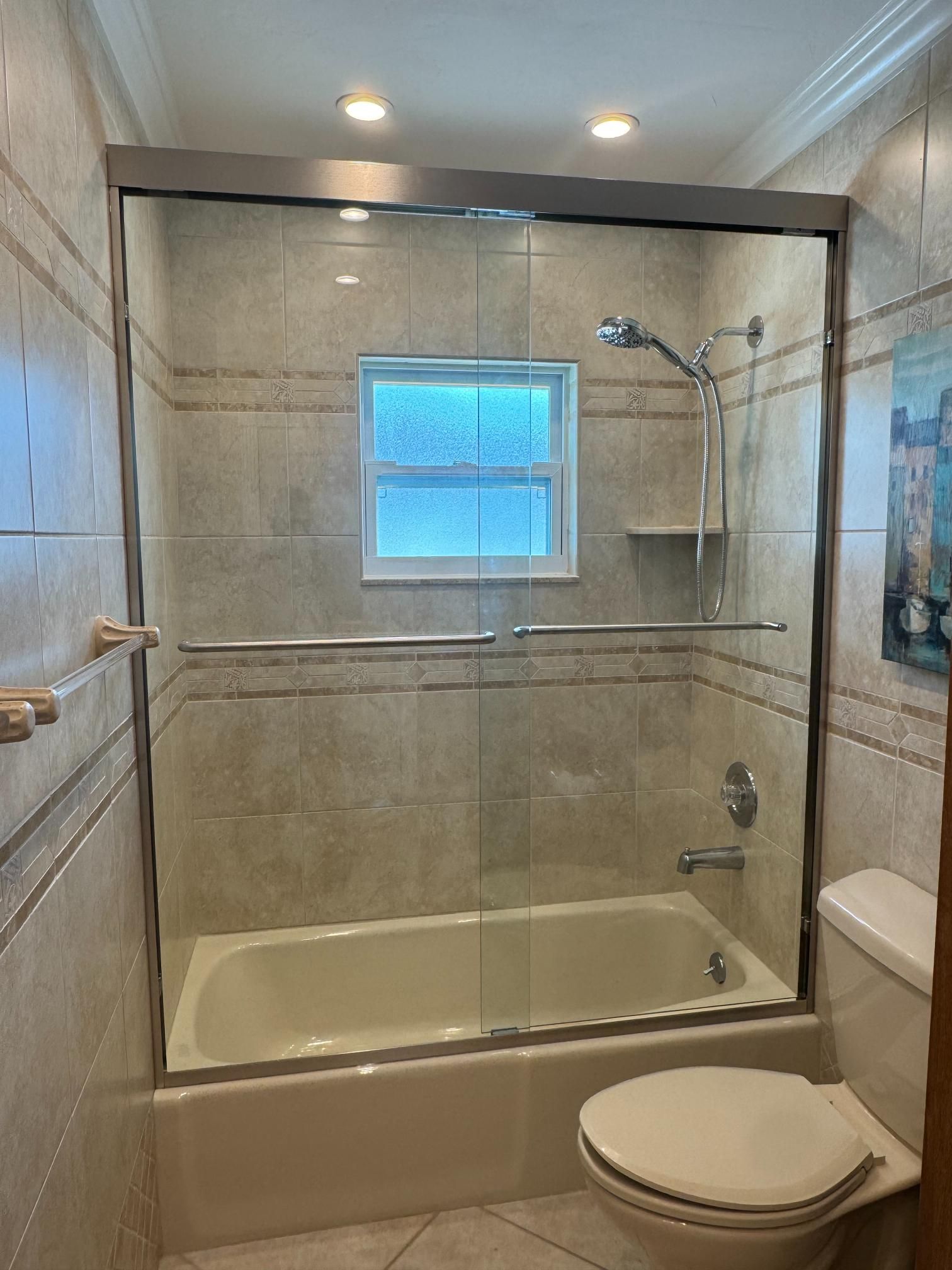 Bathroom with a bathtub, shower, toilet, and window, enclosed by glass doors and tiled walls.