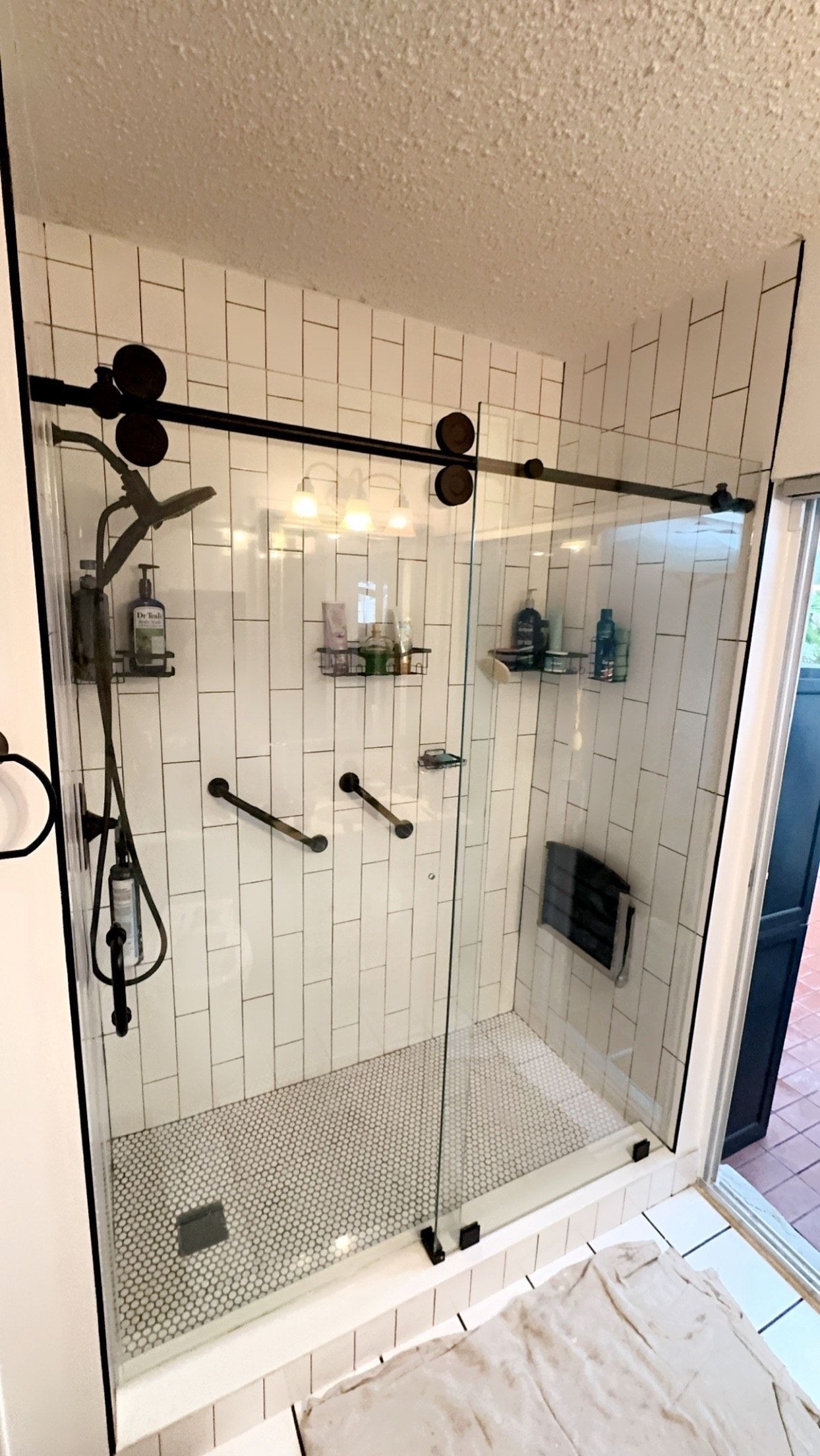 Modern white tiled shower with black accents, glass door, grab bars, and toiletries.