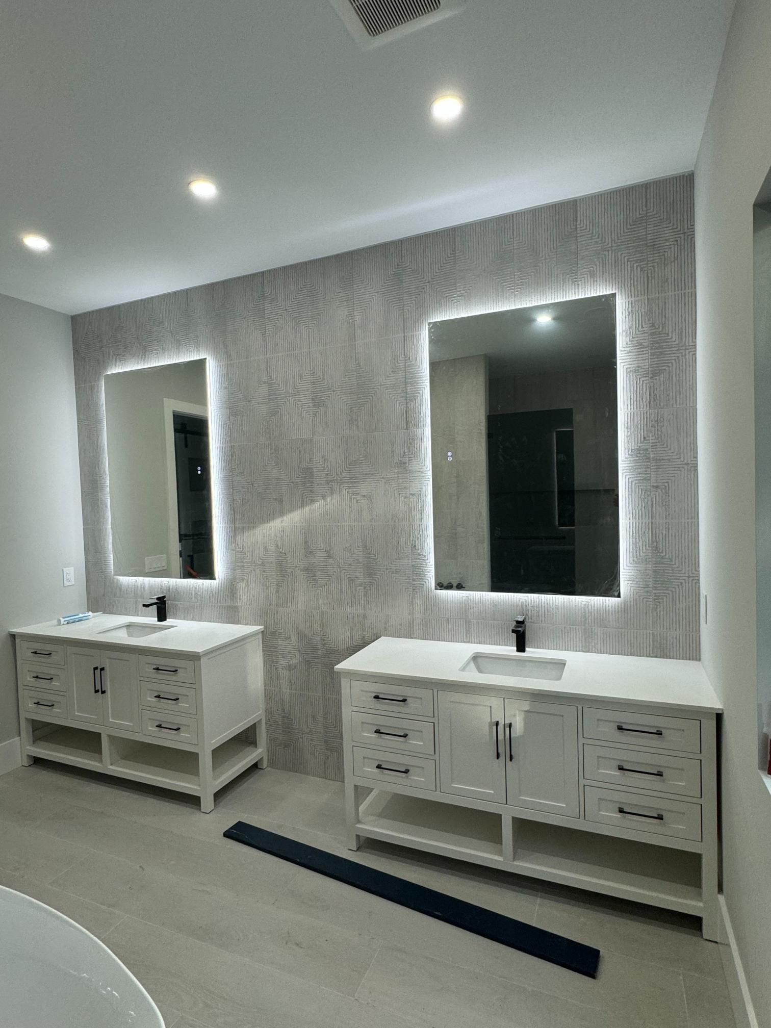 Two white bathroom vanities with mirrors and overhead lighting against a textured gray wall.