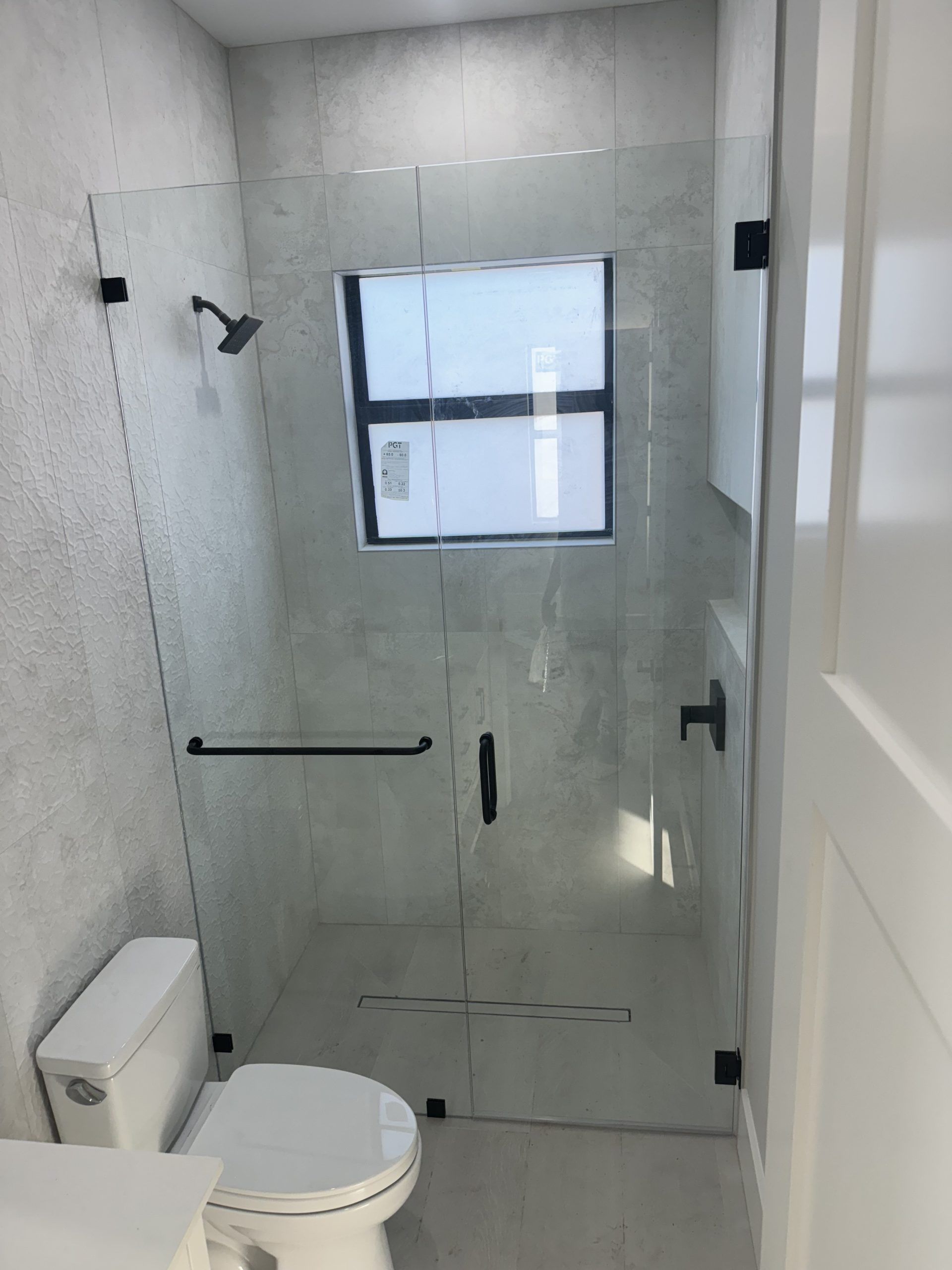 Bathroom with glass shower door, black hardware, toilet, and window. Gray tiled walls and white door.