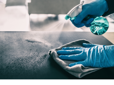 A person wearing blue gloves is cleaning a table with a cloth and spray bottle.