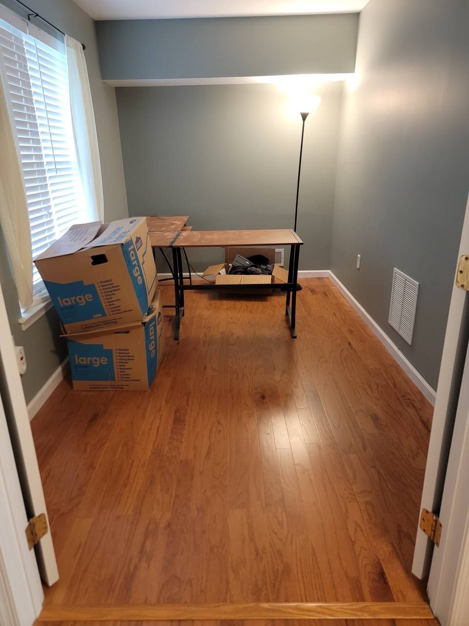 A newly painted room with a new hardwood floor, with boxes on the floor and a desk.