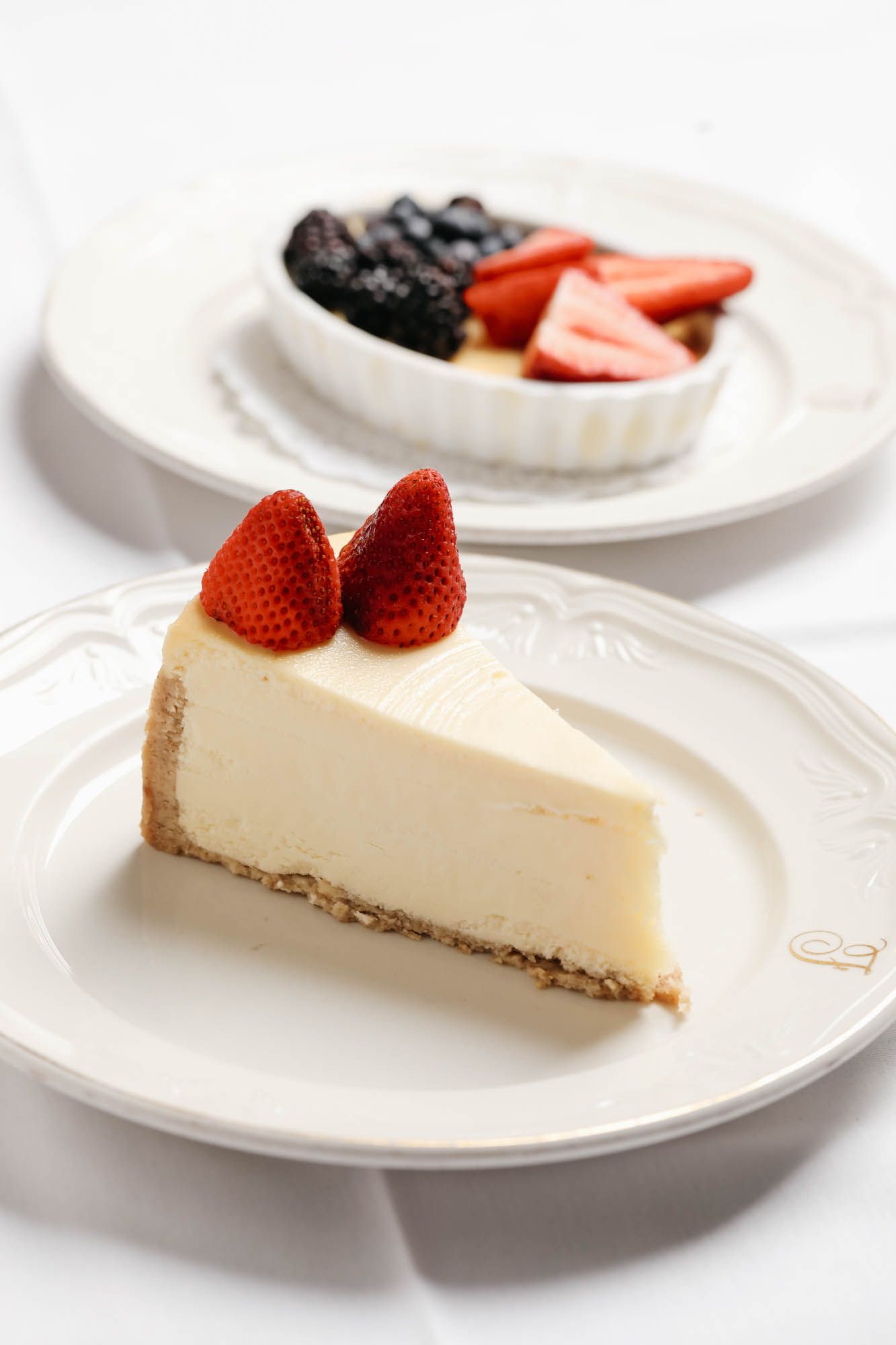A slice of cheesecake with strawberries on top is on a plate next to a bowl of fruit.