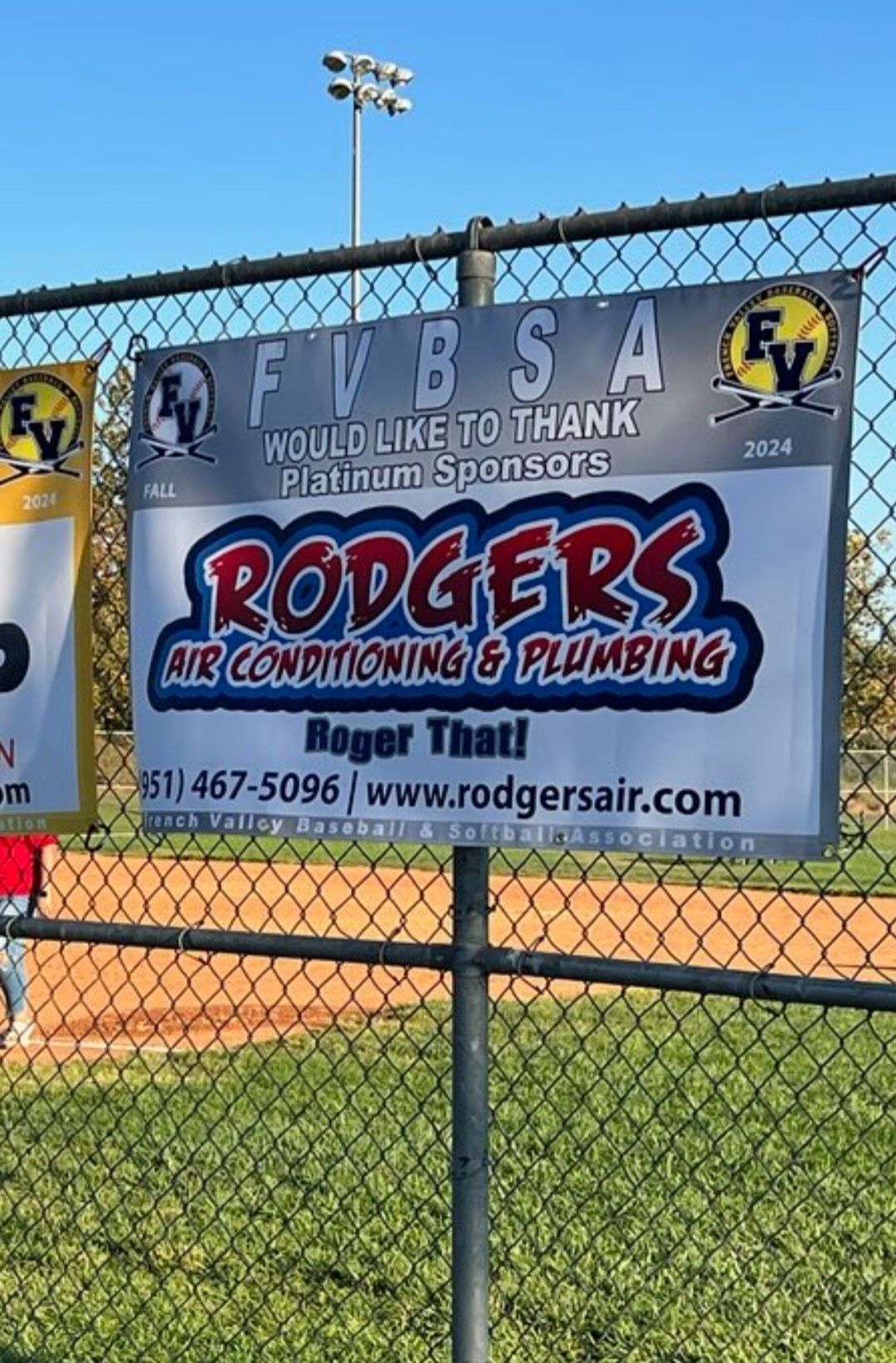 A rodgers air conditioning and plumbing sign is hanging on a chain link fence.
