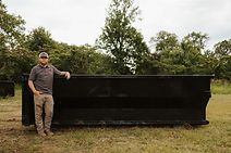 A man is standing next to a large dumpster in a field.
