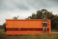 A man is standing next to an orange dumpster in a field.
