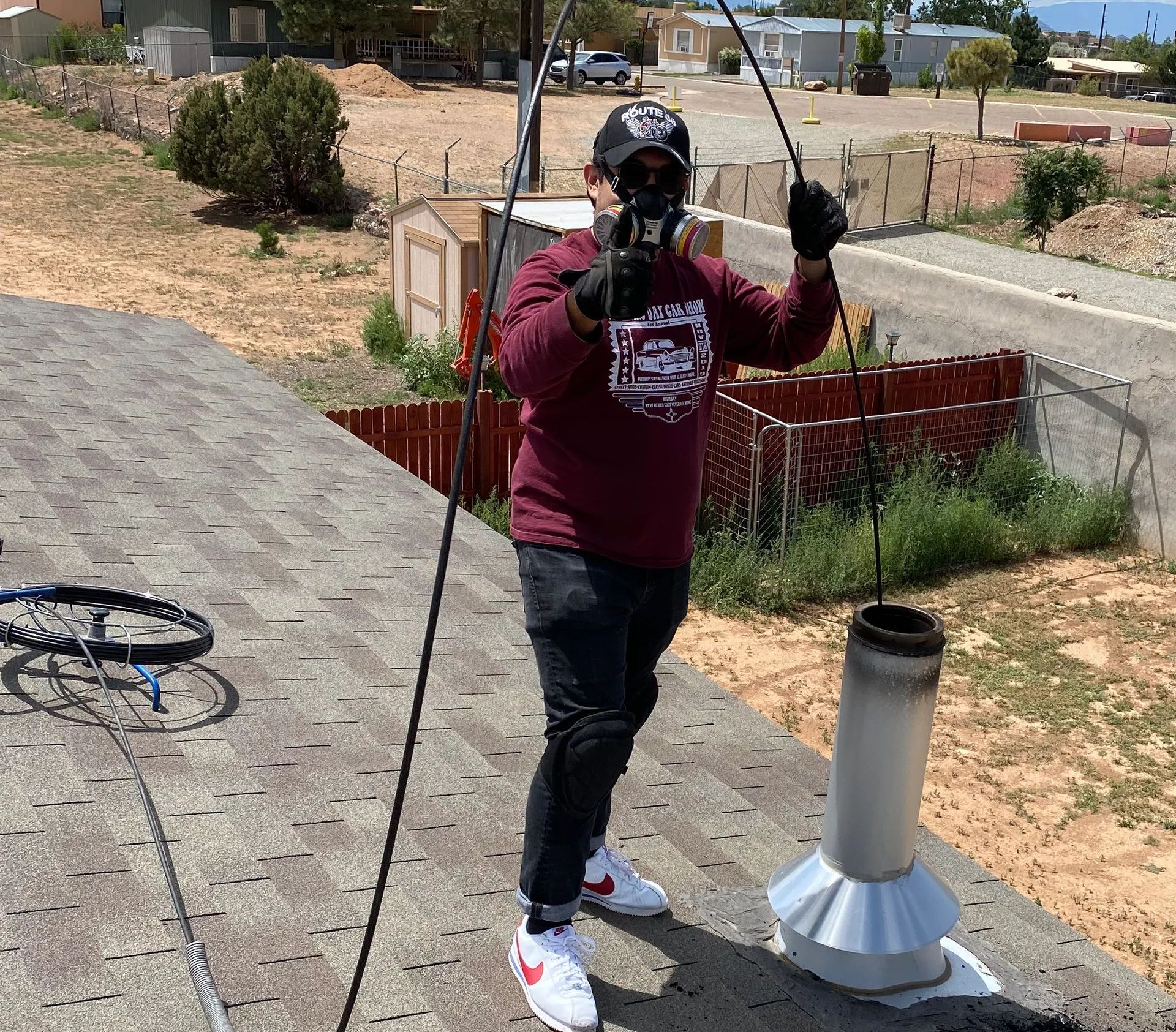 Person on a roof cleaning a chimney, wearing a mask, gloves, and red shirt, with tools and a view of houses.