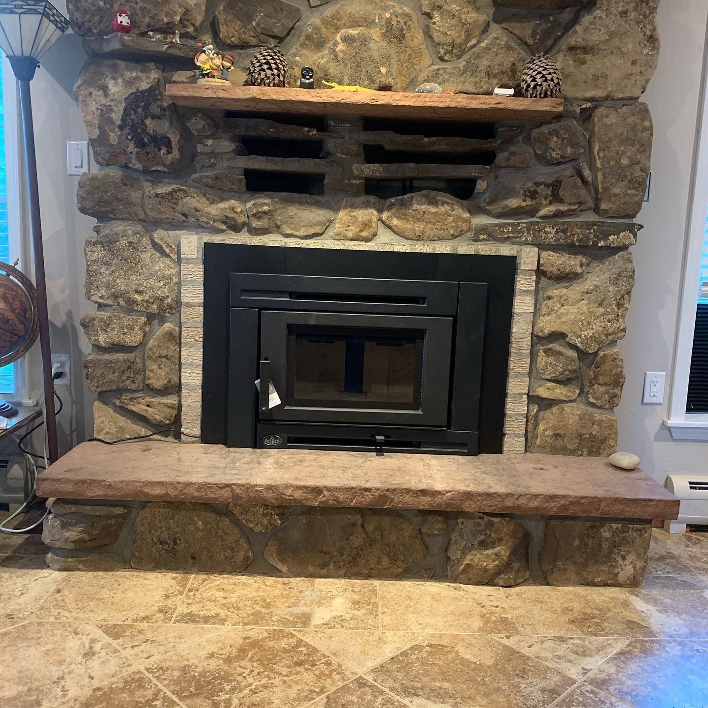 A black fireplace insert set in a rustic stone wall with a stone hearth and wooden mantel in a room with tiled floors.