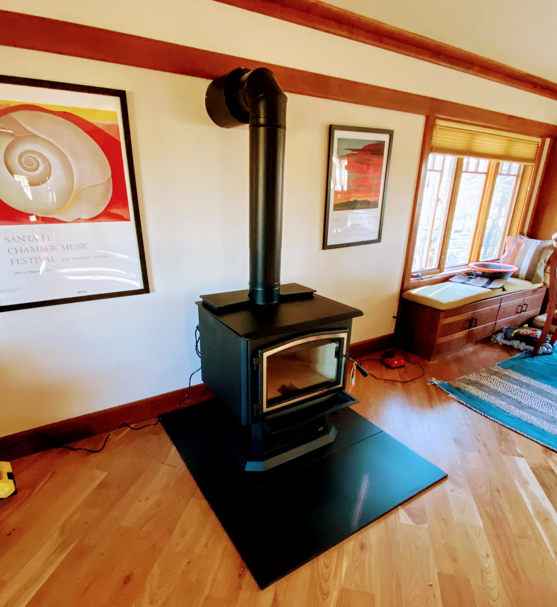 Black wood stove with a black chimney pipe on a black floor plate, in a room with hardwood floors and artwork.