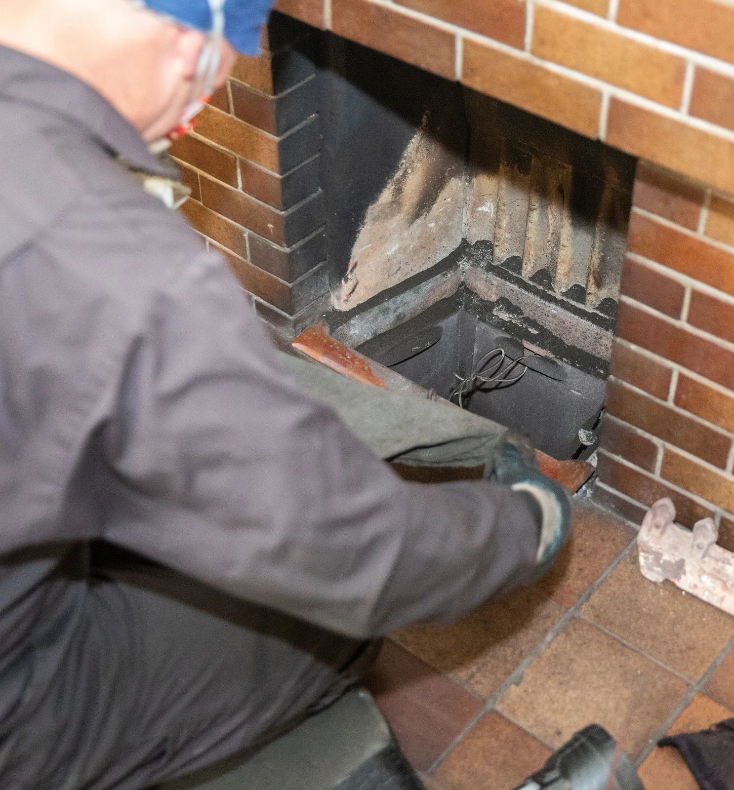 Person in grey coveralls cleaning a fireplace, holding a debris-catching piece. Brick surround, tile floor.
