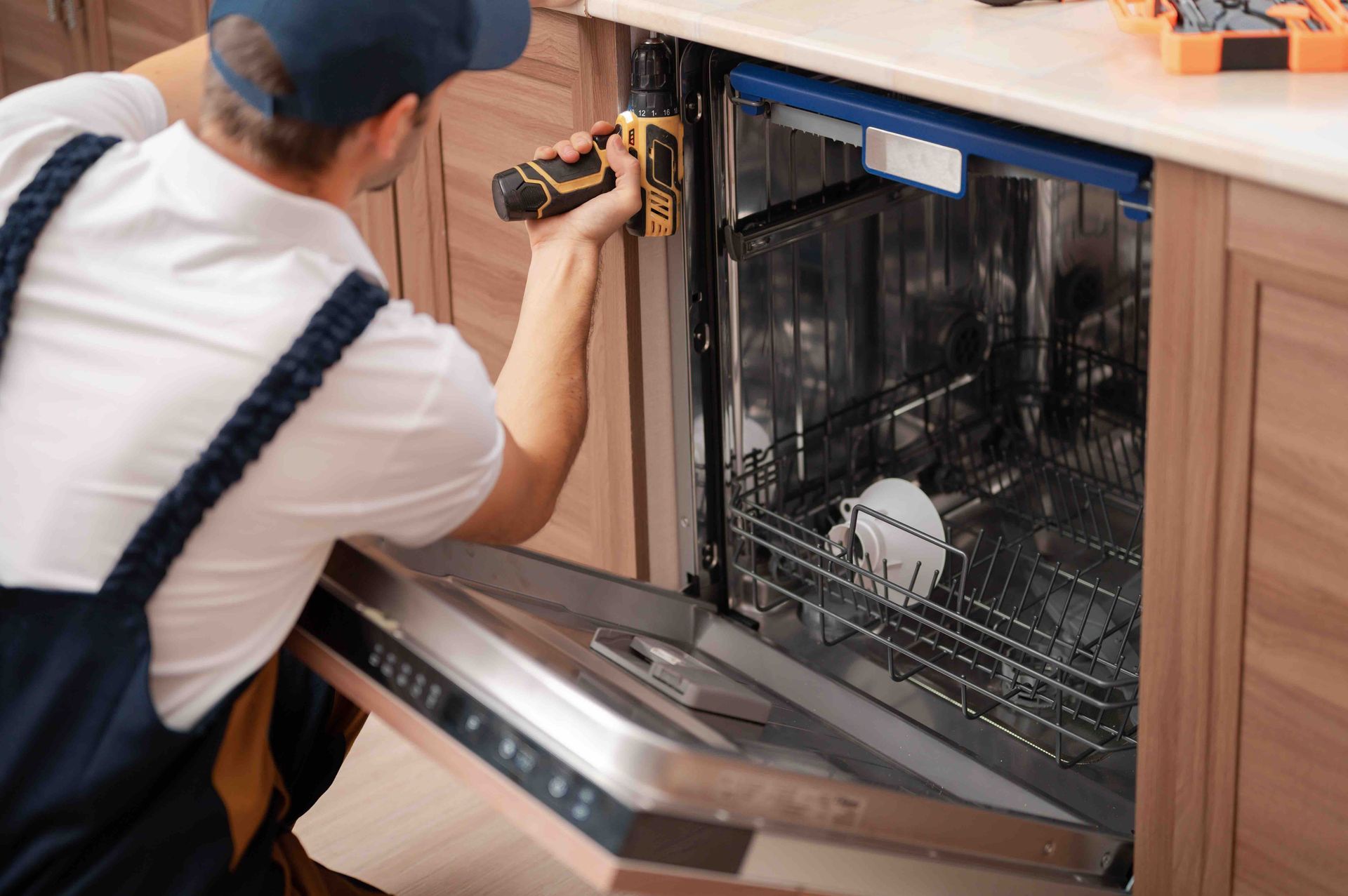 A man is fixing a dishwasher in a kitchen with a drill.
