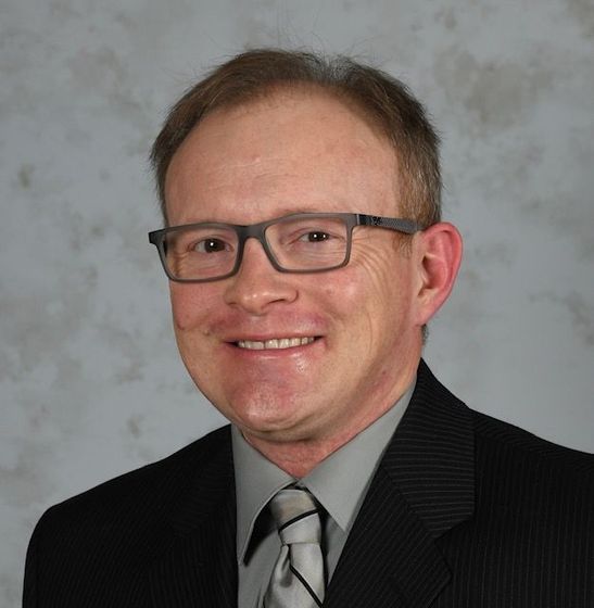 Man with glasses in a suit smiles. Background is a blurred mix of gray and white.