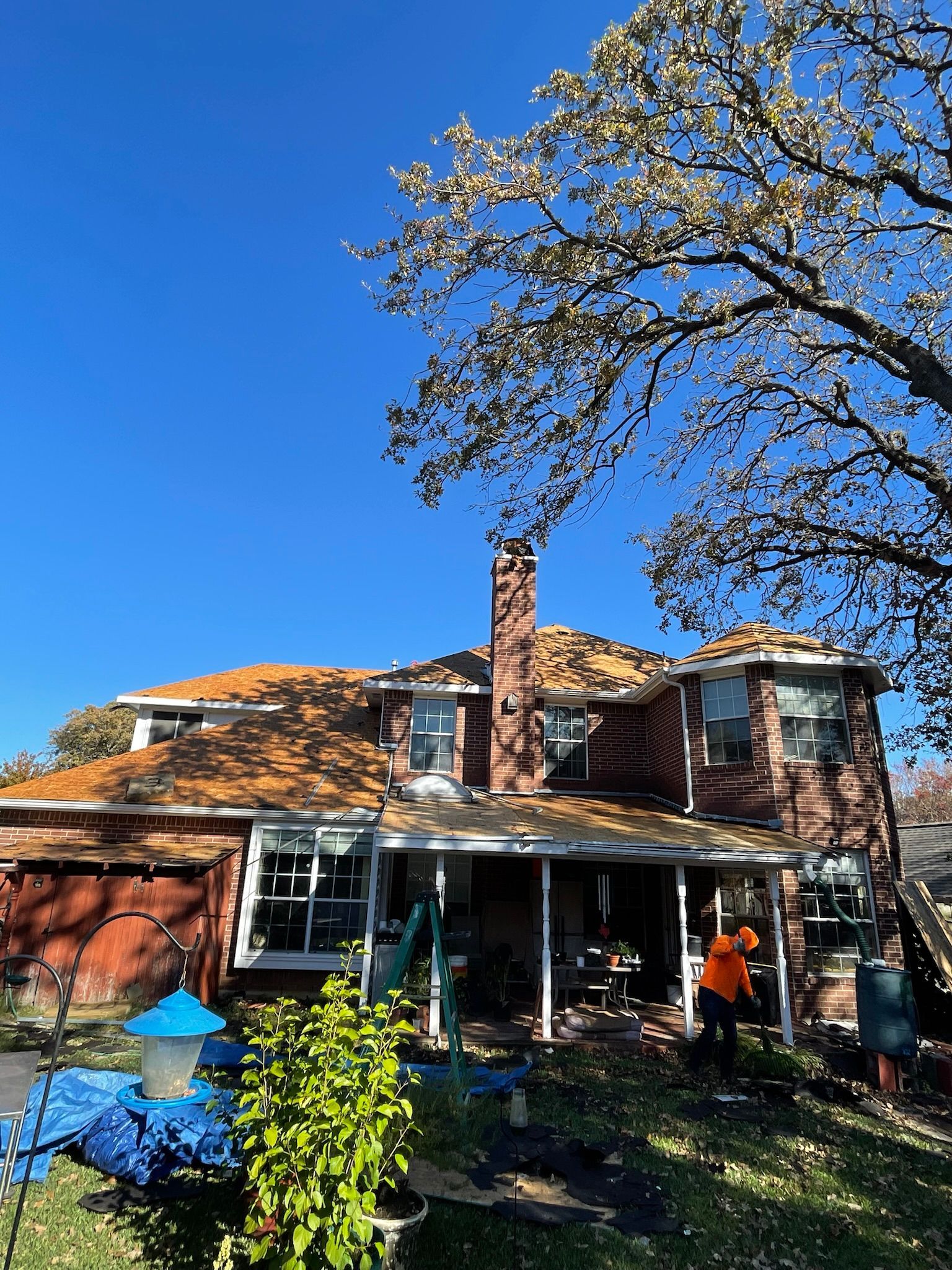 House with roof being worked on; brick chimney, blue tarp, fallen leaves, sunny day.