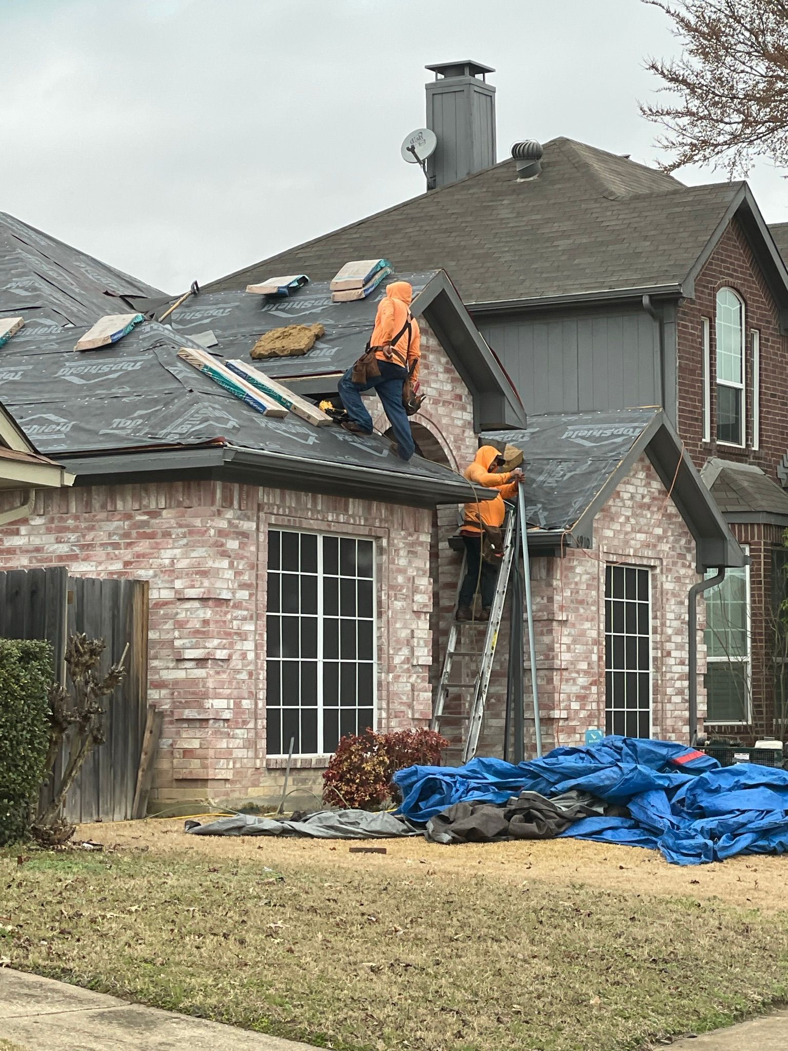 Two roofers in orange vests working on a house roof. Bricked house, blue tarp on ground.