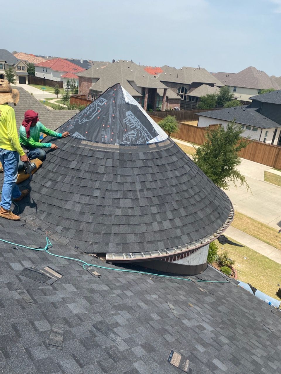 Two roofers installing shingles on a conical roof section on a sunny day.