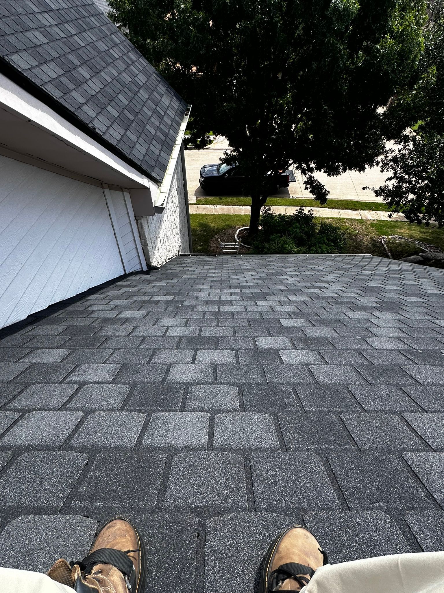 Person on a roof, looking down at the street. Dark gray shingles, white garage, green tree, and a car in the distance.