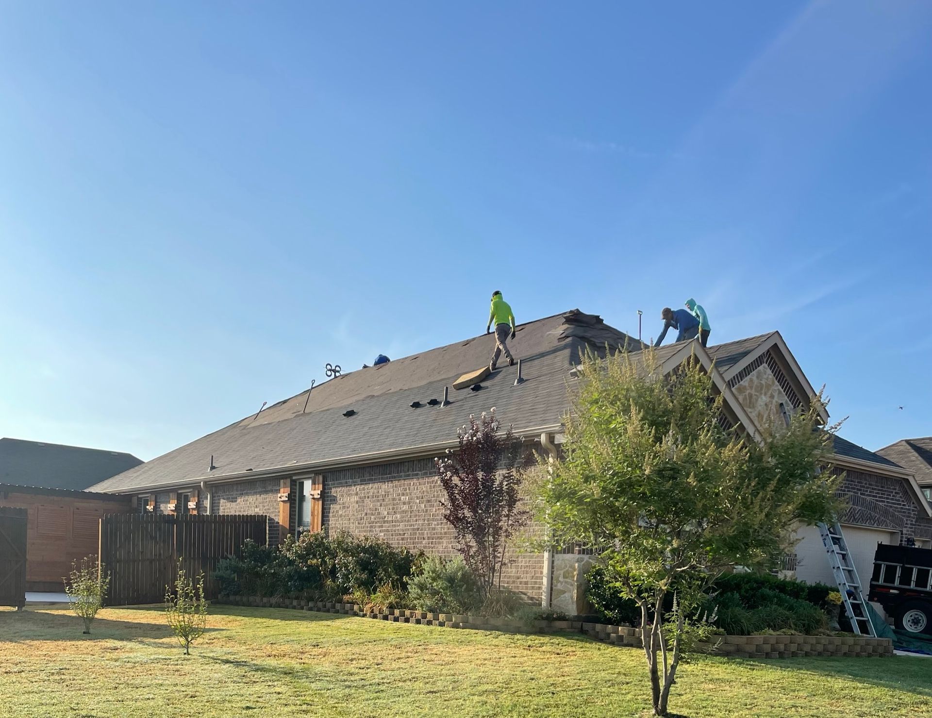 Roofers working on a dark shingled roof under a clear blue sky. A ladder leans against the house.