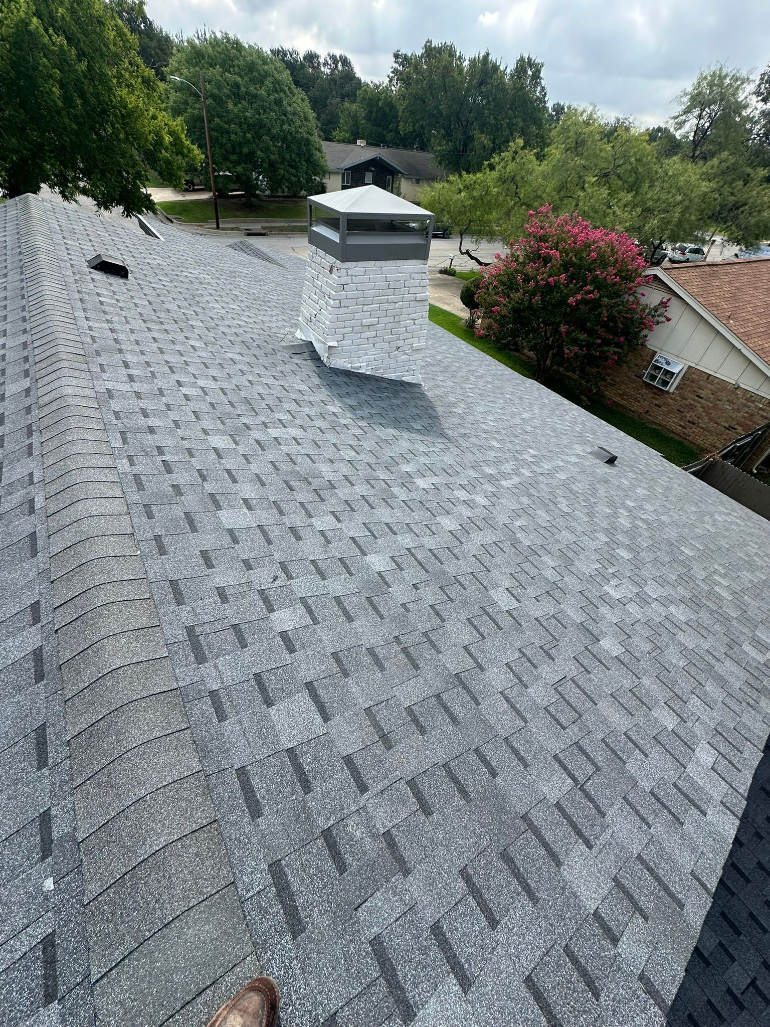 Gray asphalt shingle roof with a chimney, surrounded by trees and other houses.