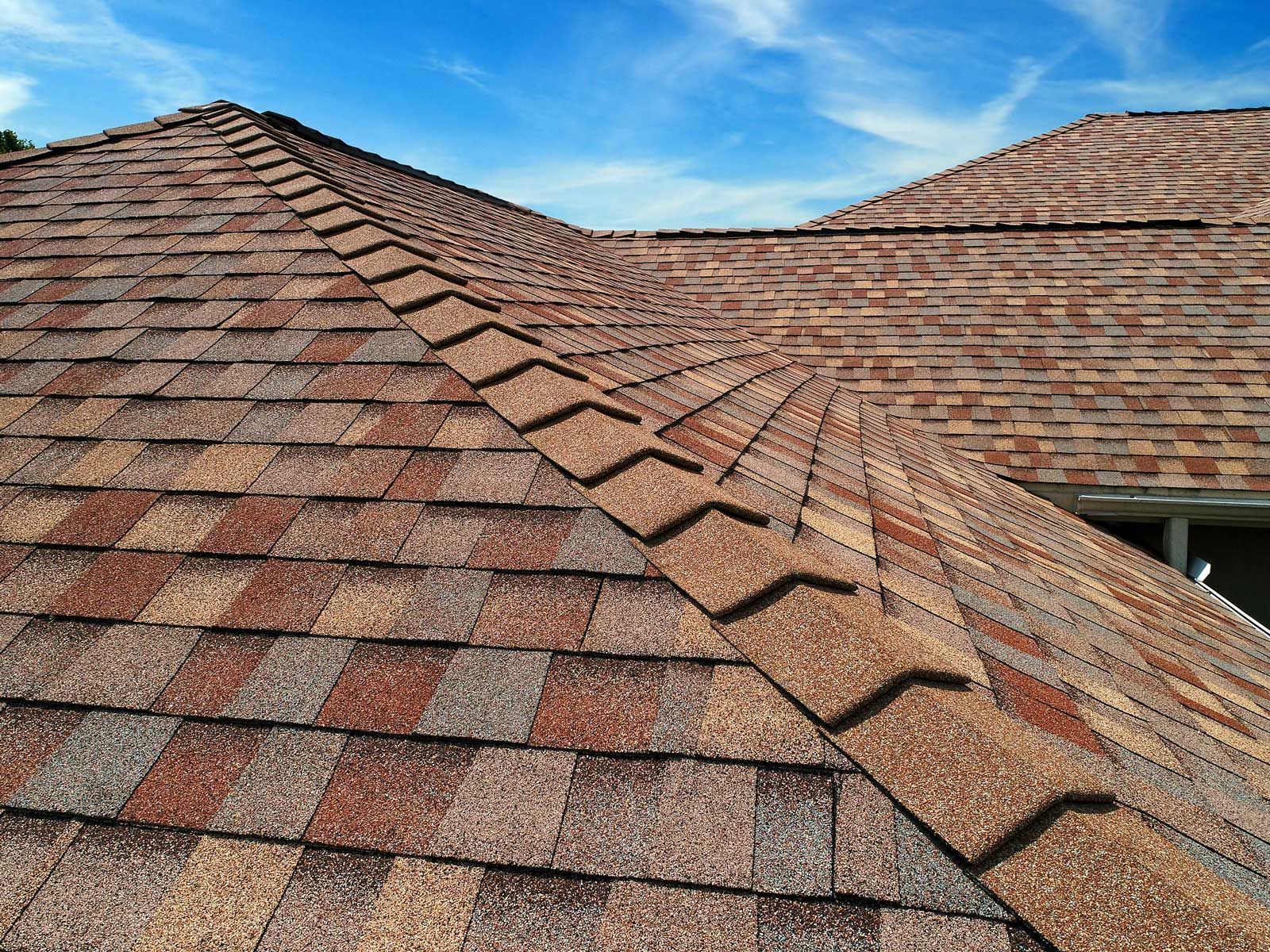 Brown and red asphalt shingle roof with a ridge cap against a blue sky.
