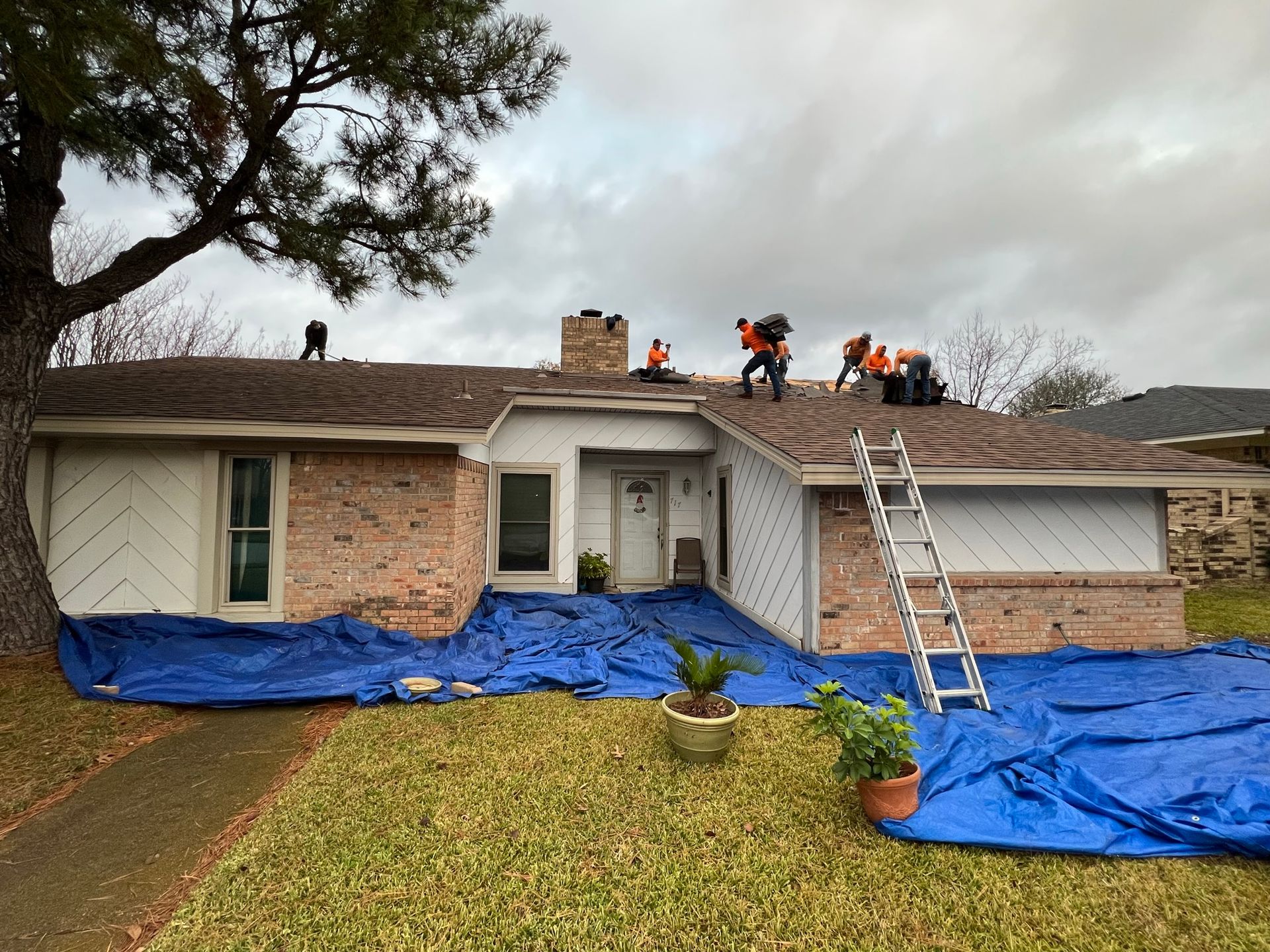 Roofing crew working on a house with brick exterior, covered with blue tarps, cloudy sky.