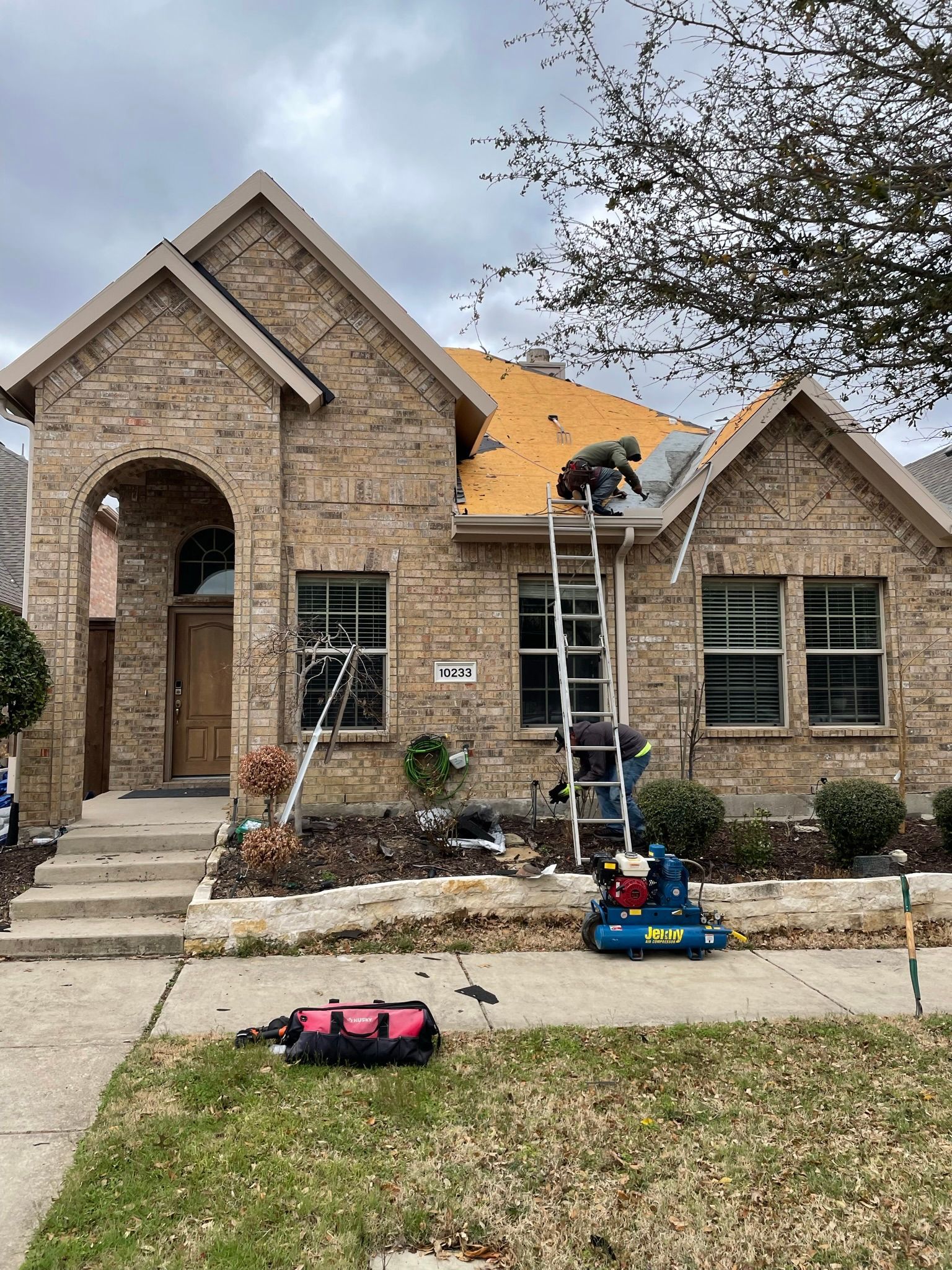 Workers repairing roof on a brick house, using a ladder. Tools on the ground. Overcast sky.