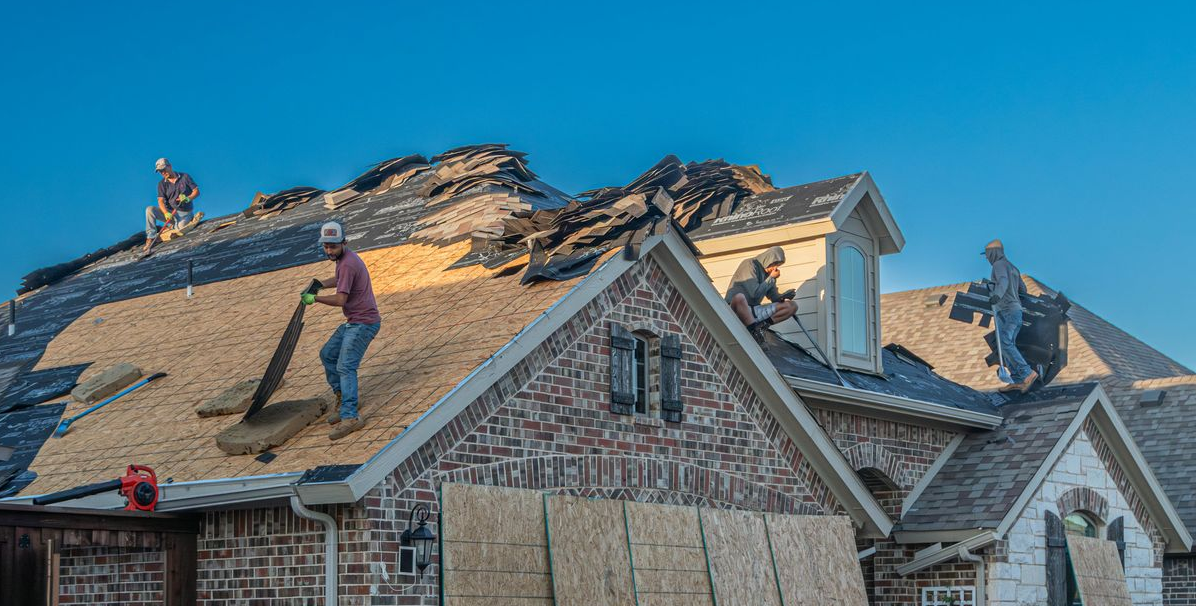 Roofers remove old shingles from a brick home's roof under a clear blue sky.